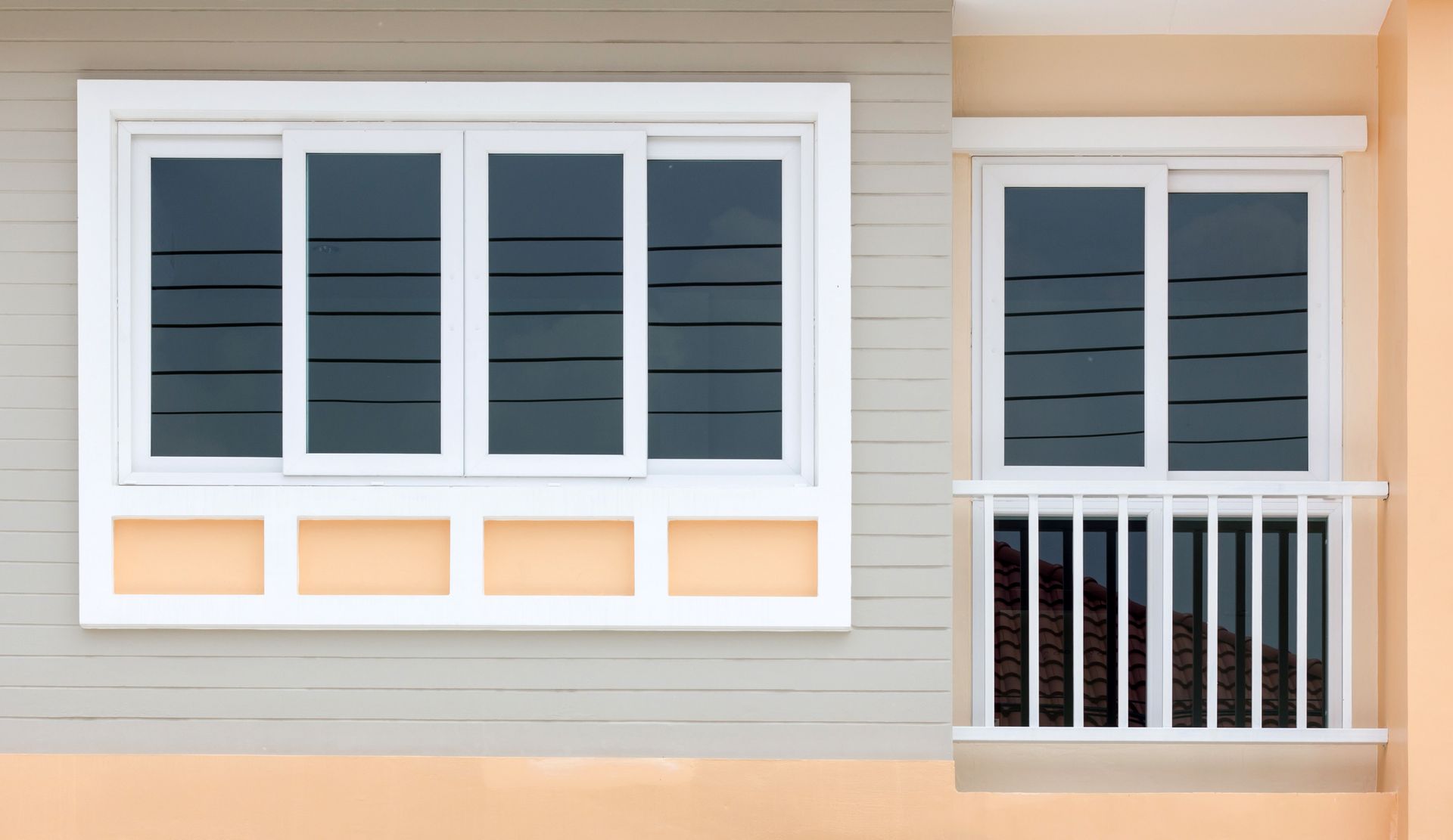 Two white-framed windows with dark panes on a beige exterior wall, one beside a small balcony railing.
