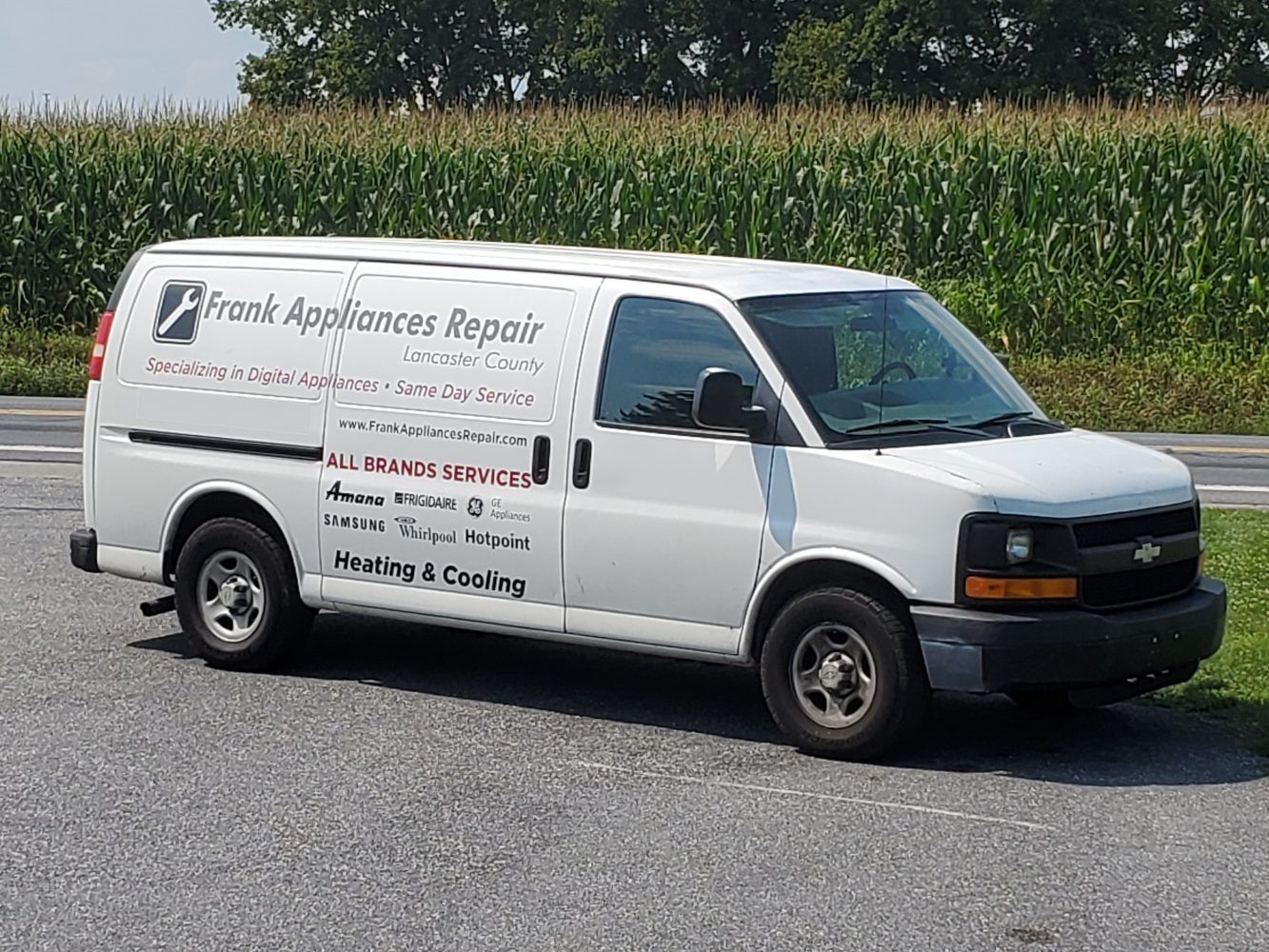 A white van is parked on the side of the road in front of a corn field.