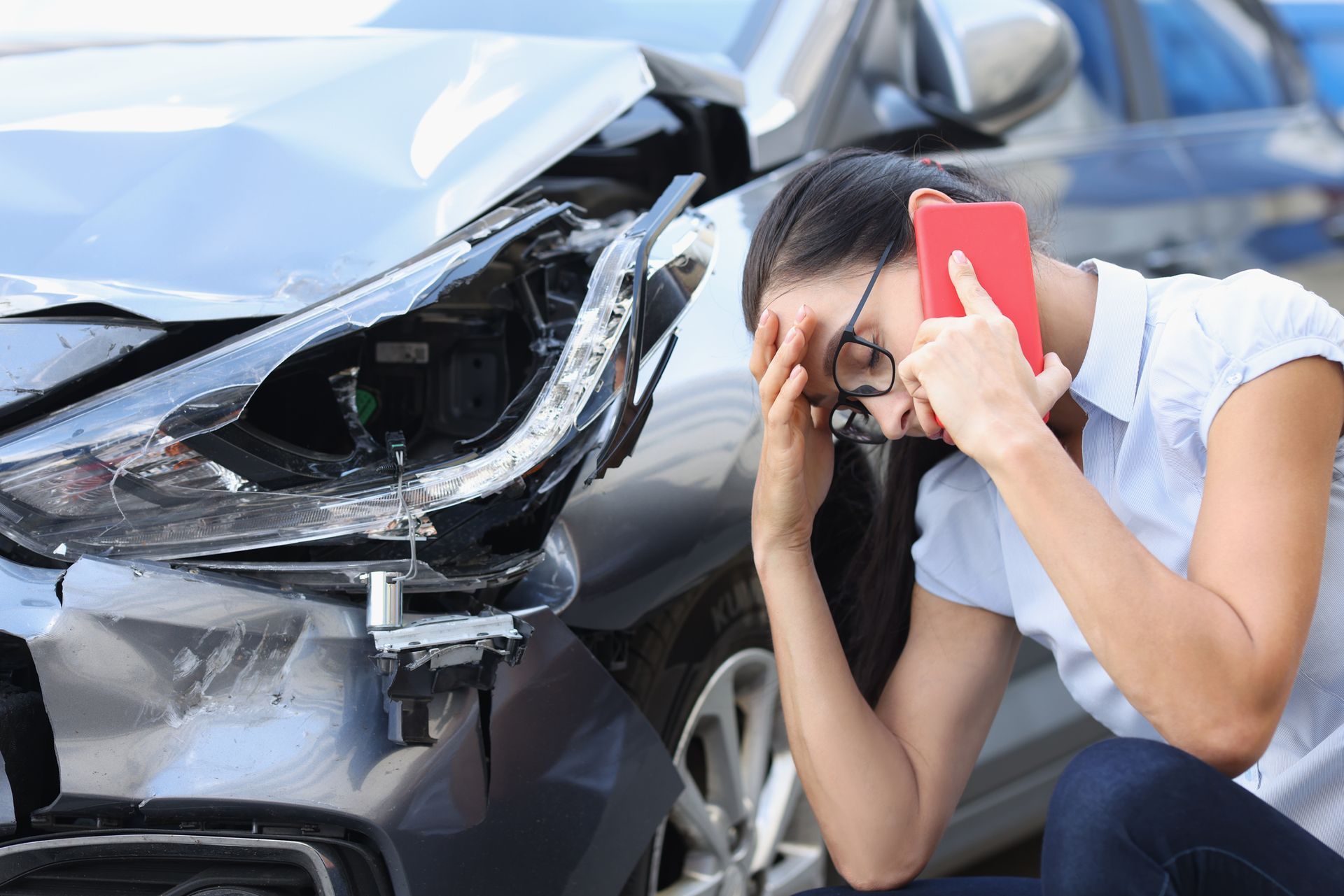 A woman is sitting in front of a damaged car talking on a cell phone.