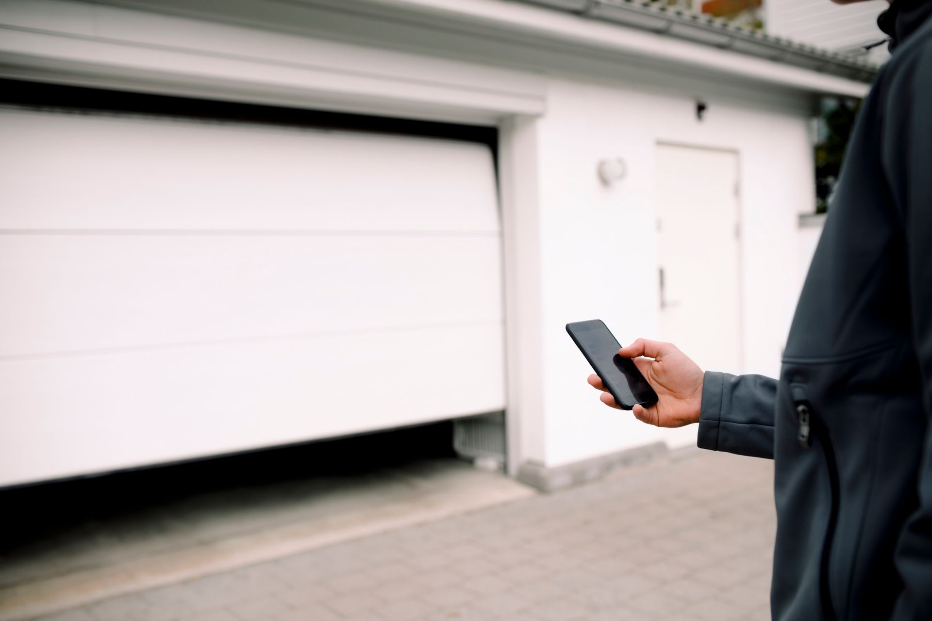 Person operating a garage door with a smartphone. The white door is partially open; a house is in the background.