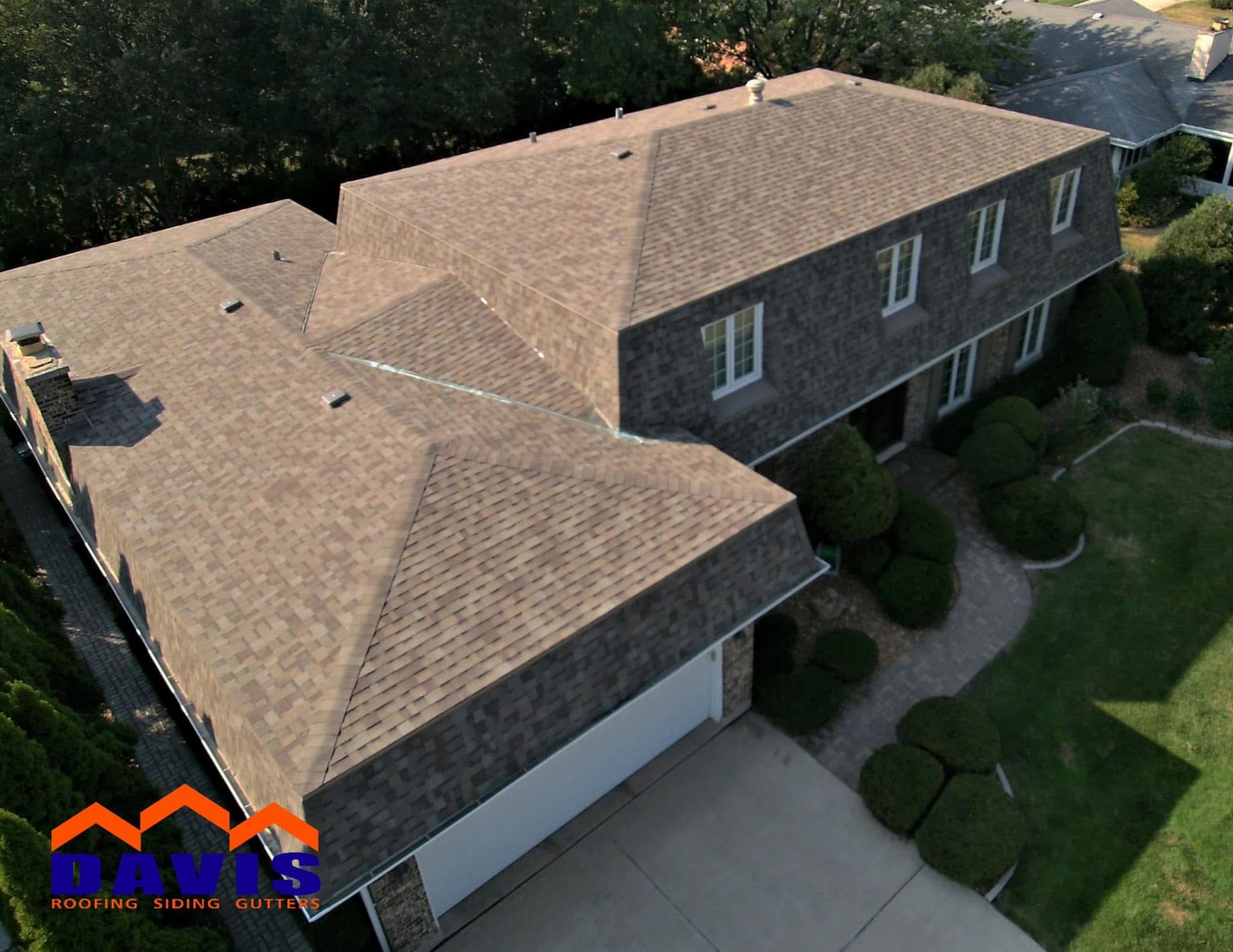 Aerial view of a brown-shingled house with multiple roof levels, a garage, and green landscaping.