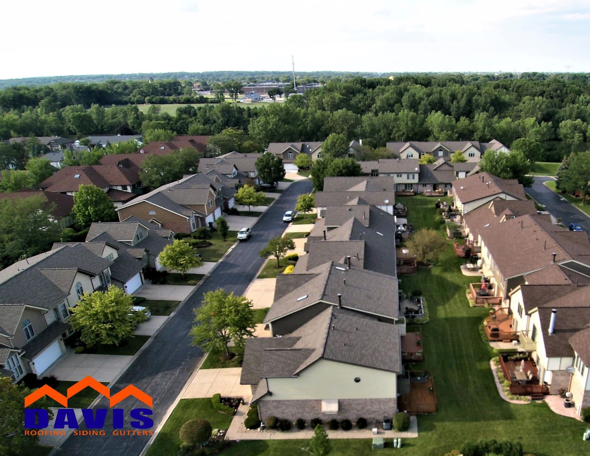Aerial view of a suburban neighborhood with houses, driveways, and green lawns, with a logo in the bottom left corner.