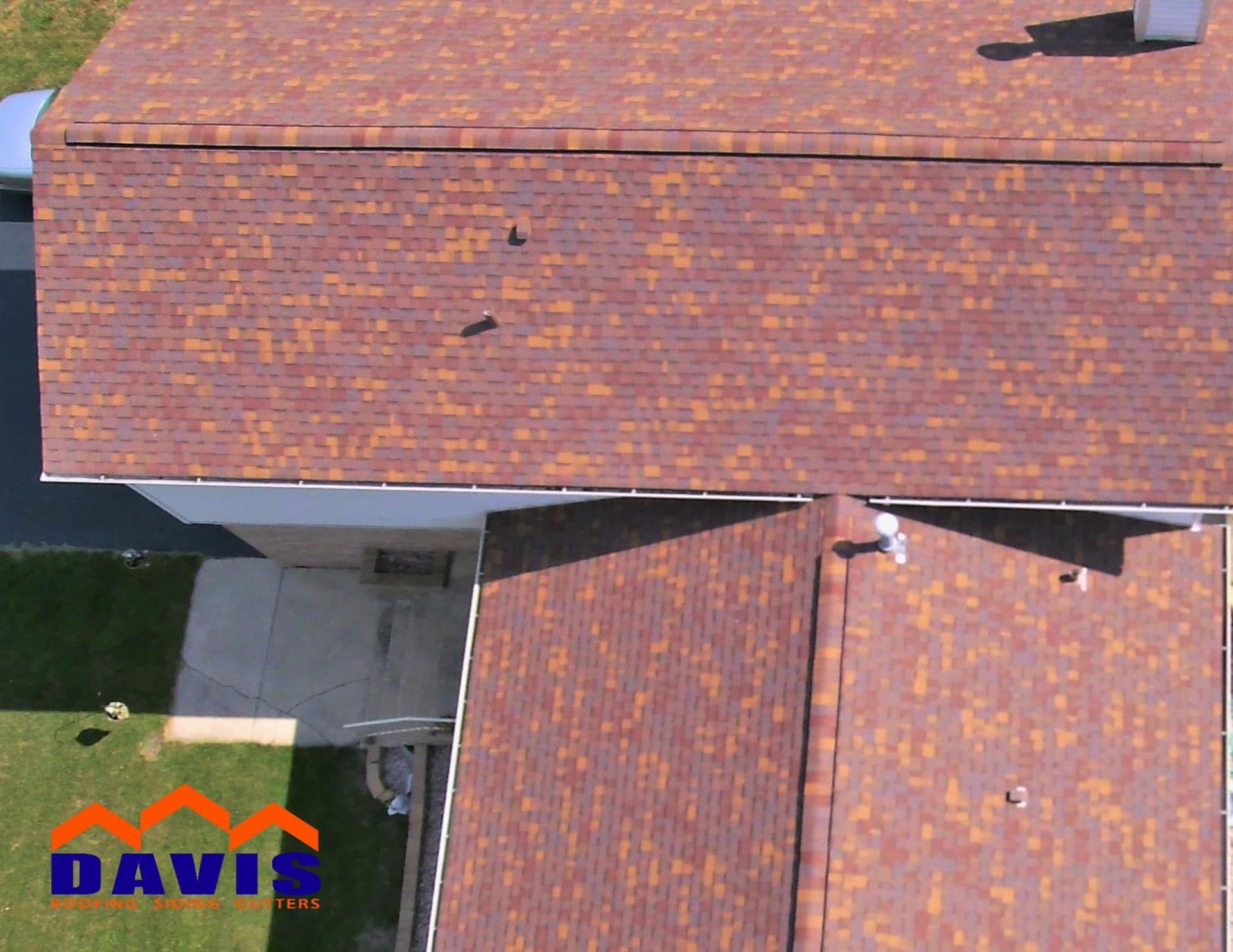 Aerial view of a brown and orange shingle roof on a house, with green grass and a concrete patio visible.