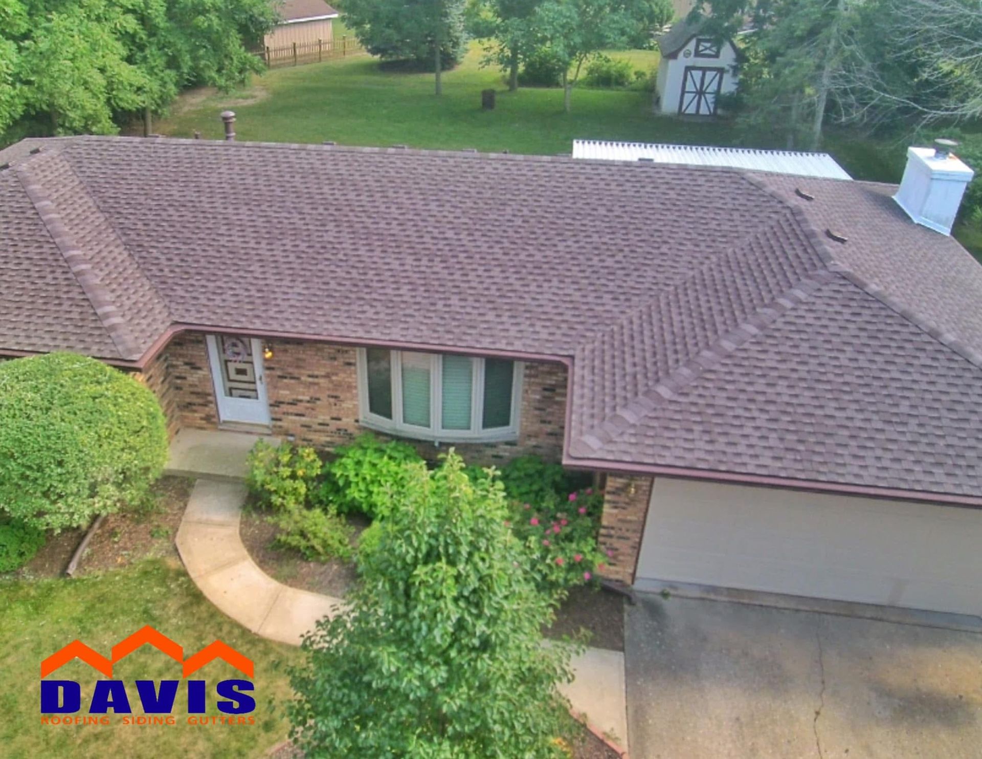 Aerial view of a brown shingle roof on a brick house with a curved walkway, trees, and green lawn.