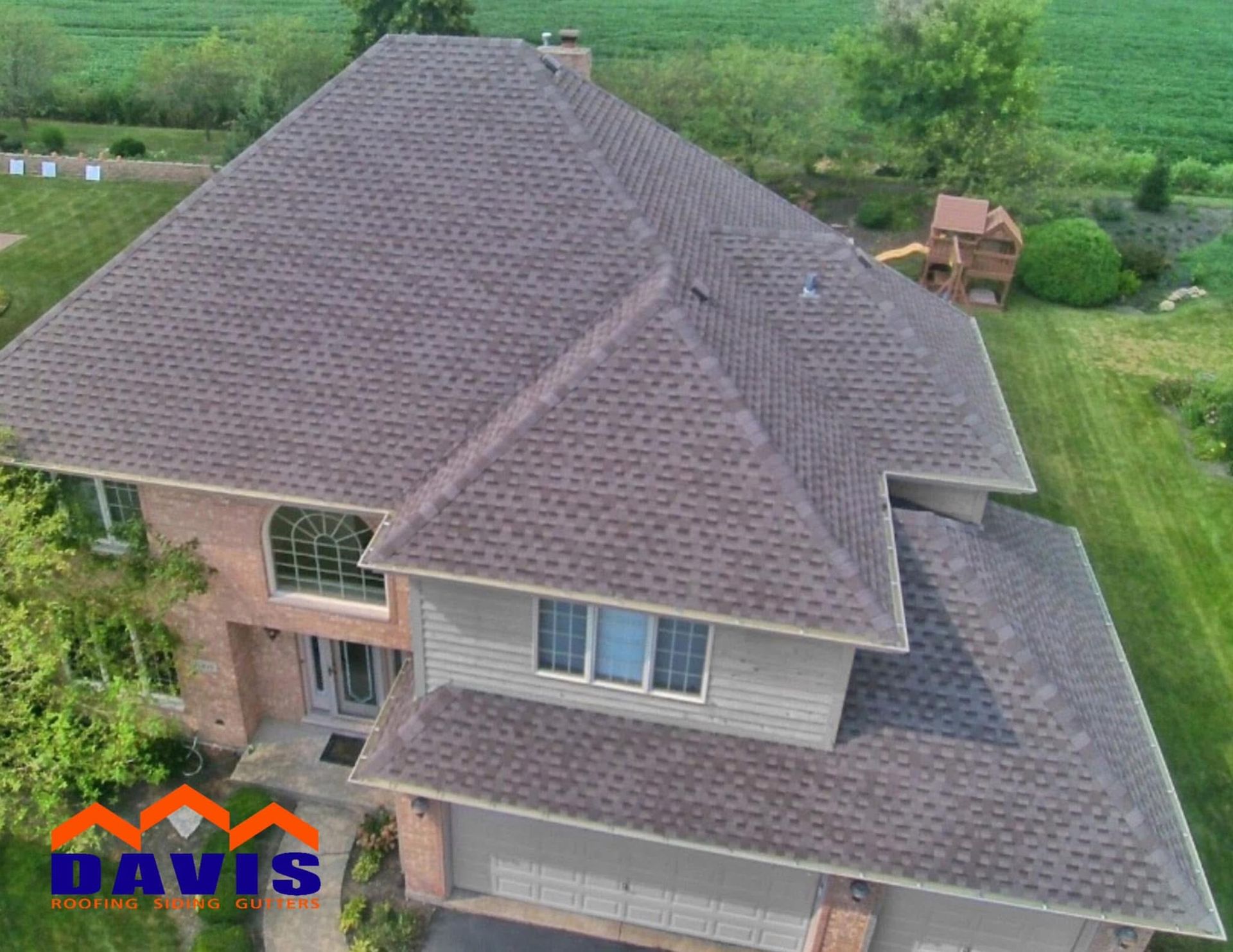 Brown shingled roof on a two-story house with tan siding and brick accents, viewed from above, surrounded by green grass.