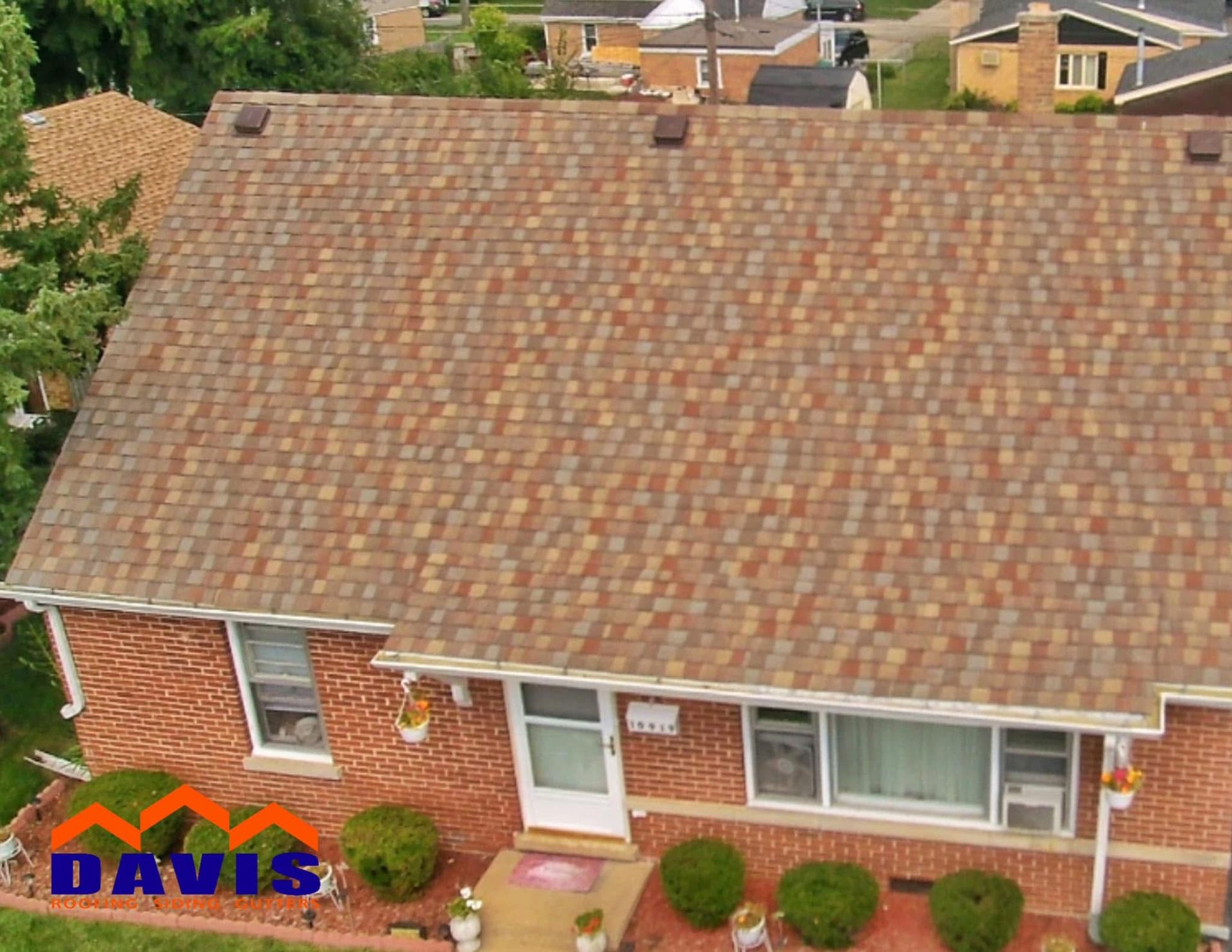 A brick home with a brown and tan shingled roof, windows, and bushes in the front yard.