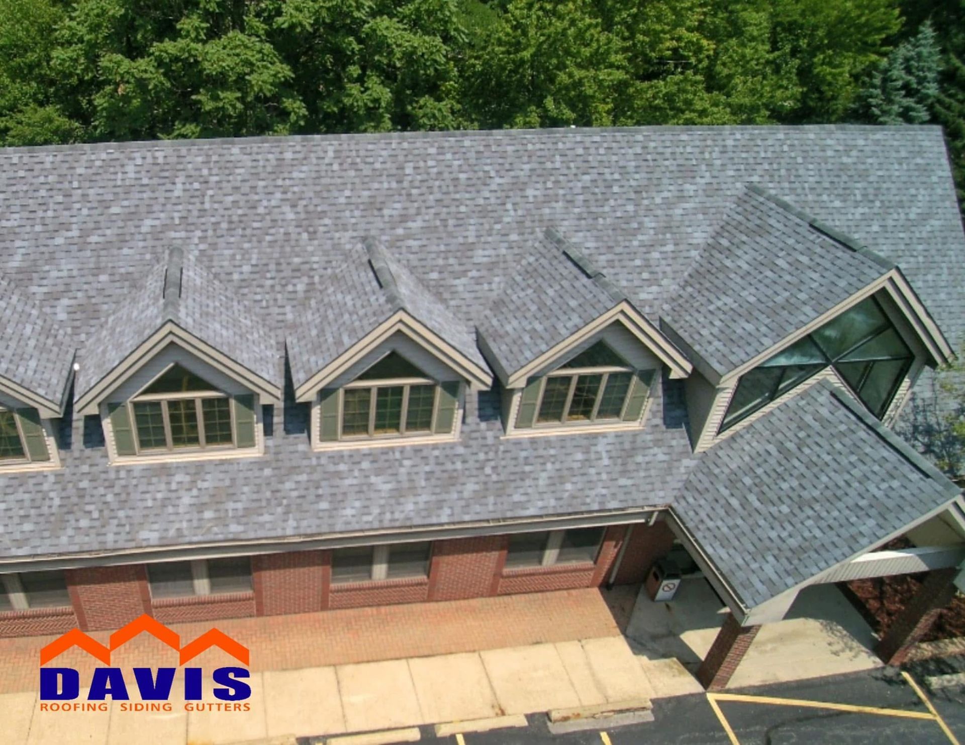 Grey shingle roof on a brick building with multiple dormers, set against a backdrop of trees.
