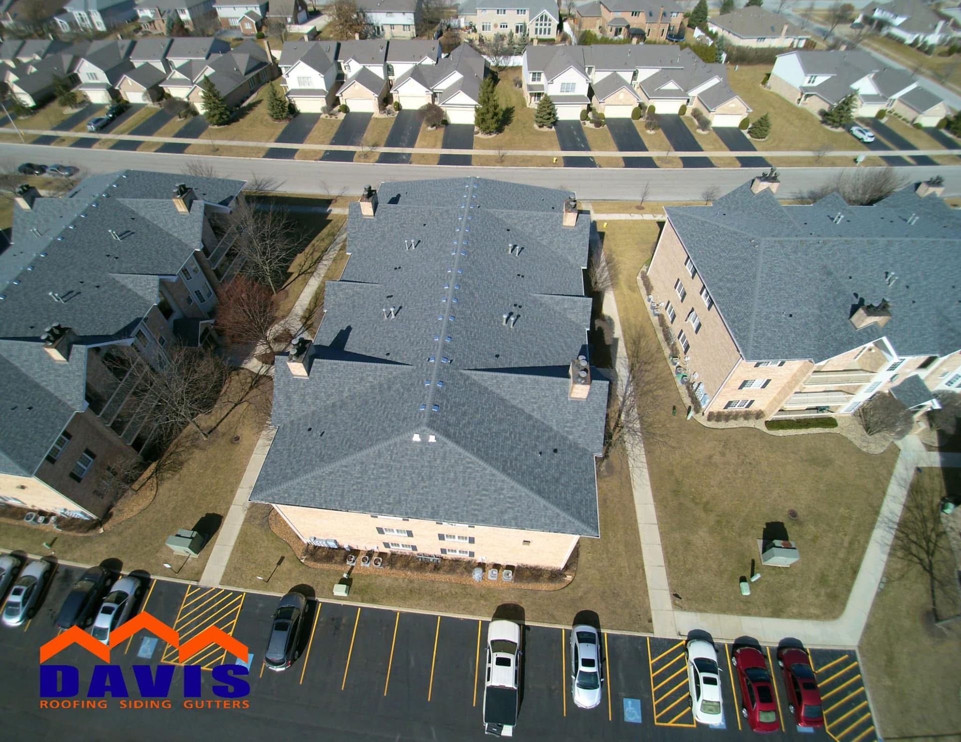 Aerial view of a complex with newly shingled roofs. Cars are parked in a lot; other homes are in the distance.