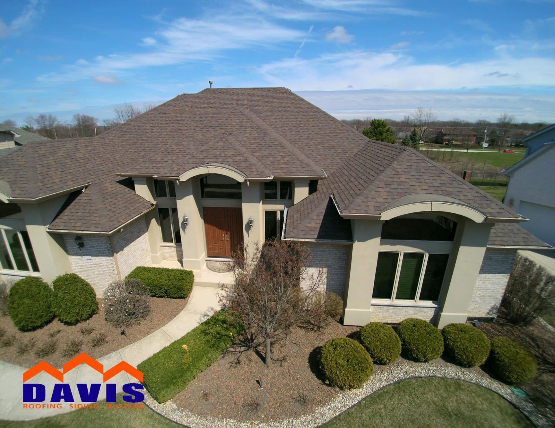 Brown-roofed house with beige walls and green landscaping under a blue sky; Davis Roofing logo in the lower left.