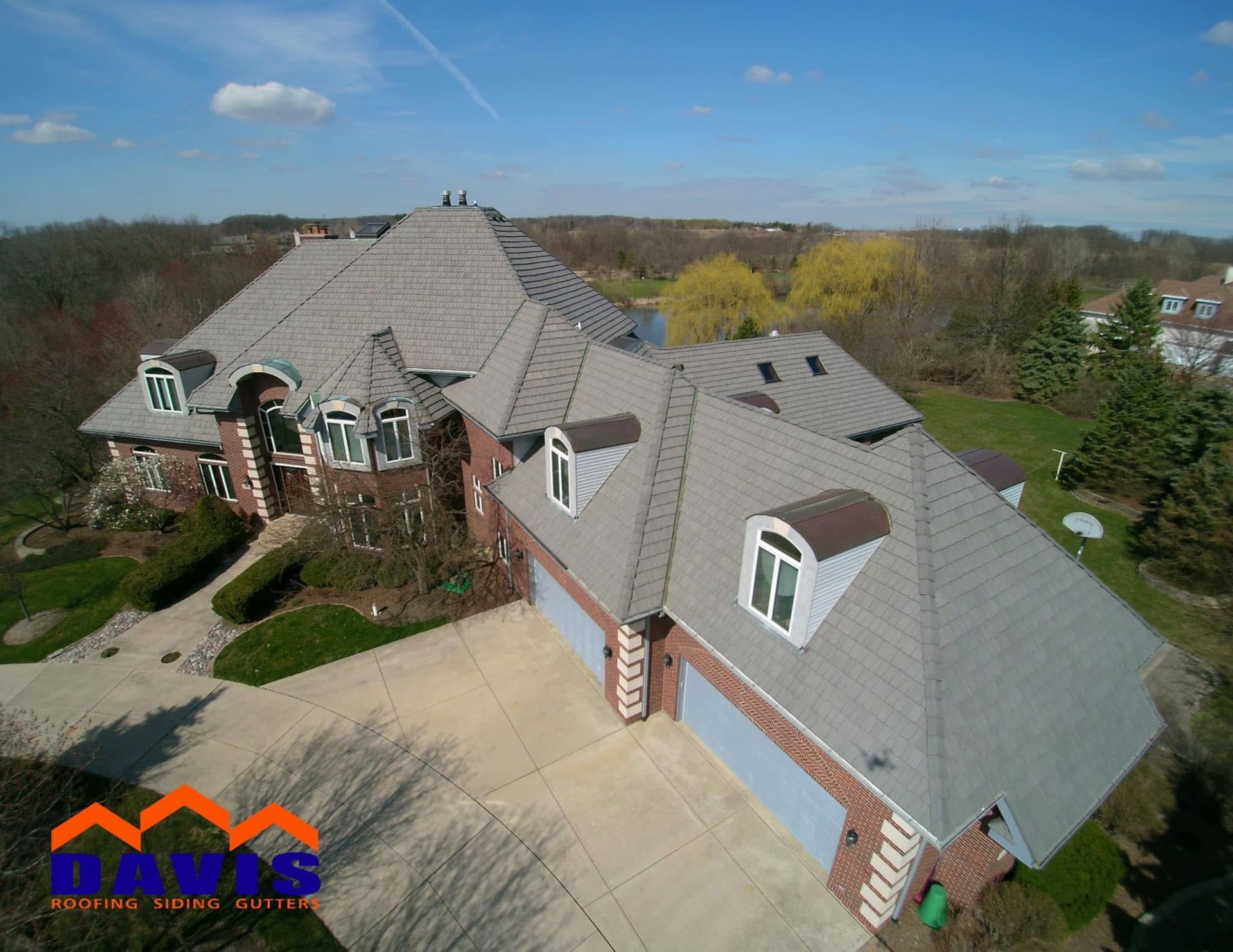Aerial view of a large home with a gray shingle roof, brick exterior, and attached garage on a sunny day.