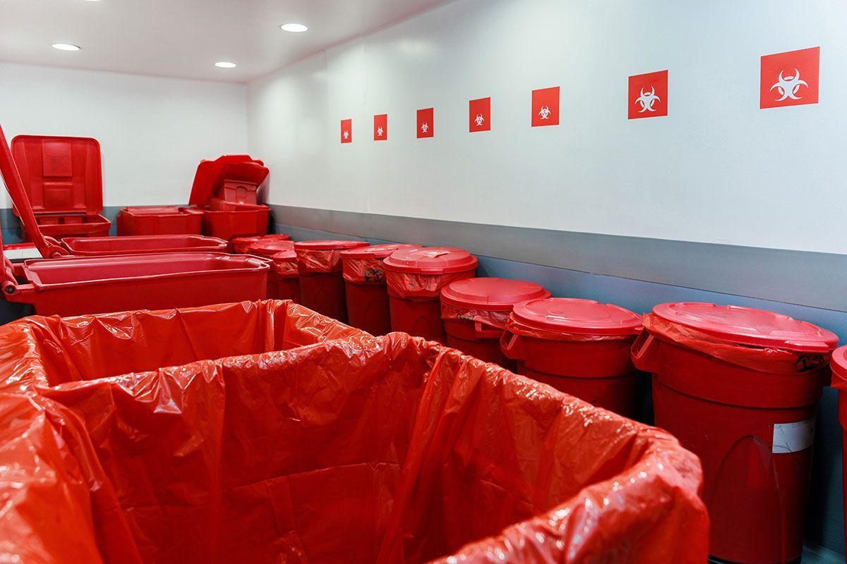 Storage room filled with red biohazard waste containers and lined red bins under biohazard warning signs on the wall.