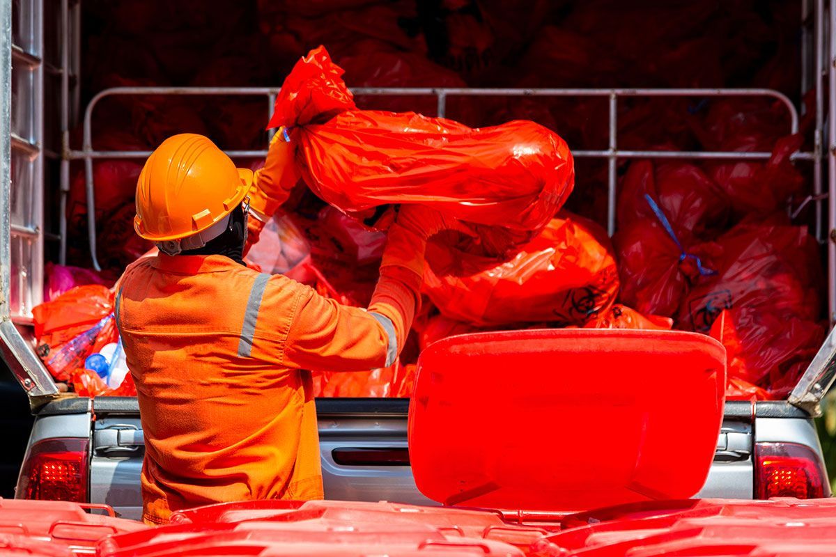 Worker in orange suit and hardhat loading red bags into a truck, possibly hazardous waste.