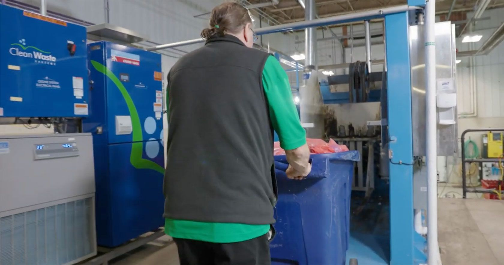 Person in green shirt and black vest using a blue waste processing machine in a facility