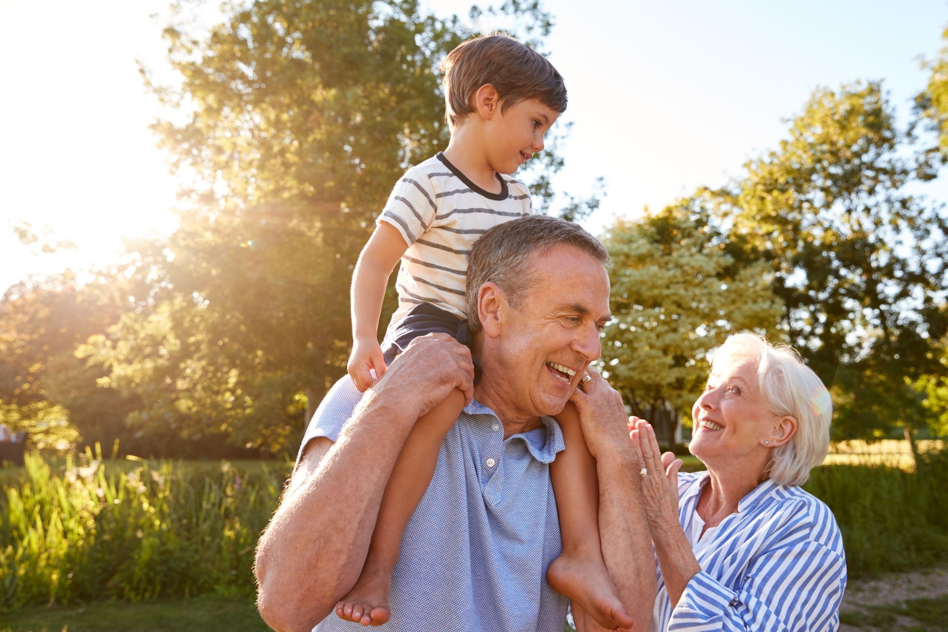 Smiling man carrying a child on his shoulders while an elderly woman smiles beside them in a sunny park
