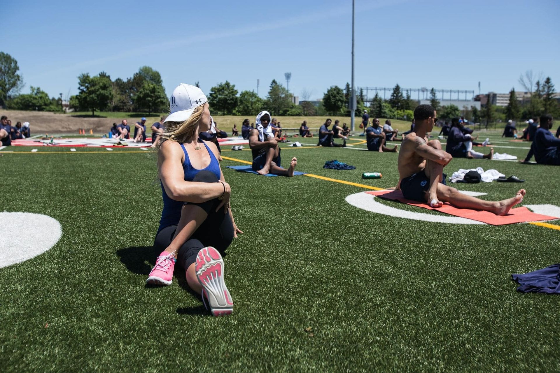 People doing yoga on a green field, sunny day. Woman in blue stretches leg, other people sit in different poses.