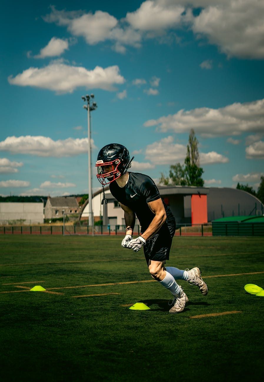 Football player running drills on a field with yellow cones under a blue sky.