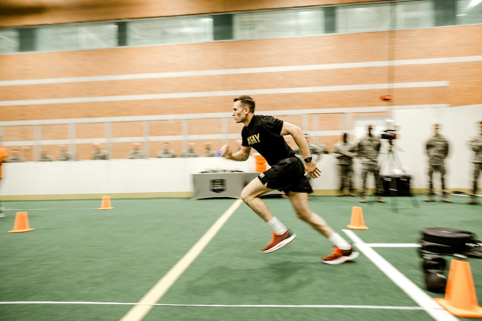 Runner sprinting indoors on a turf field with orange cones and observers in the background
