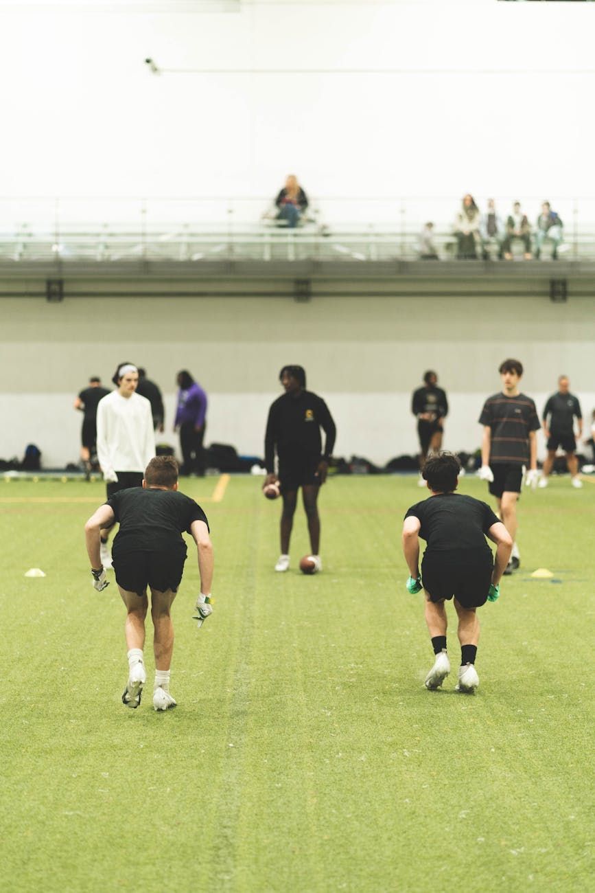 Athletes stretching on an indoor turf field, seen from behind, with spectators seated on bleachers.