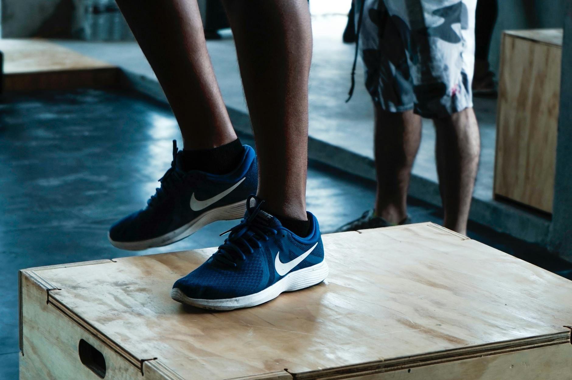 Two people standing on wooden boxes in a gym, one wearing blue sneakers.