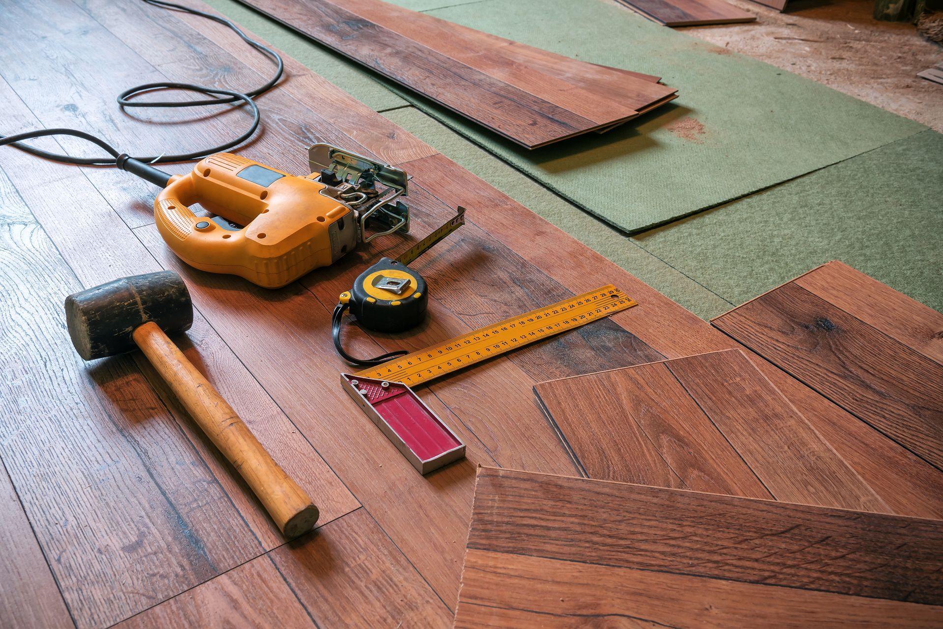 Tools and flooring materials arranged on floor: hammer, saw, measuring tape, square, wood planks.