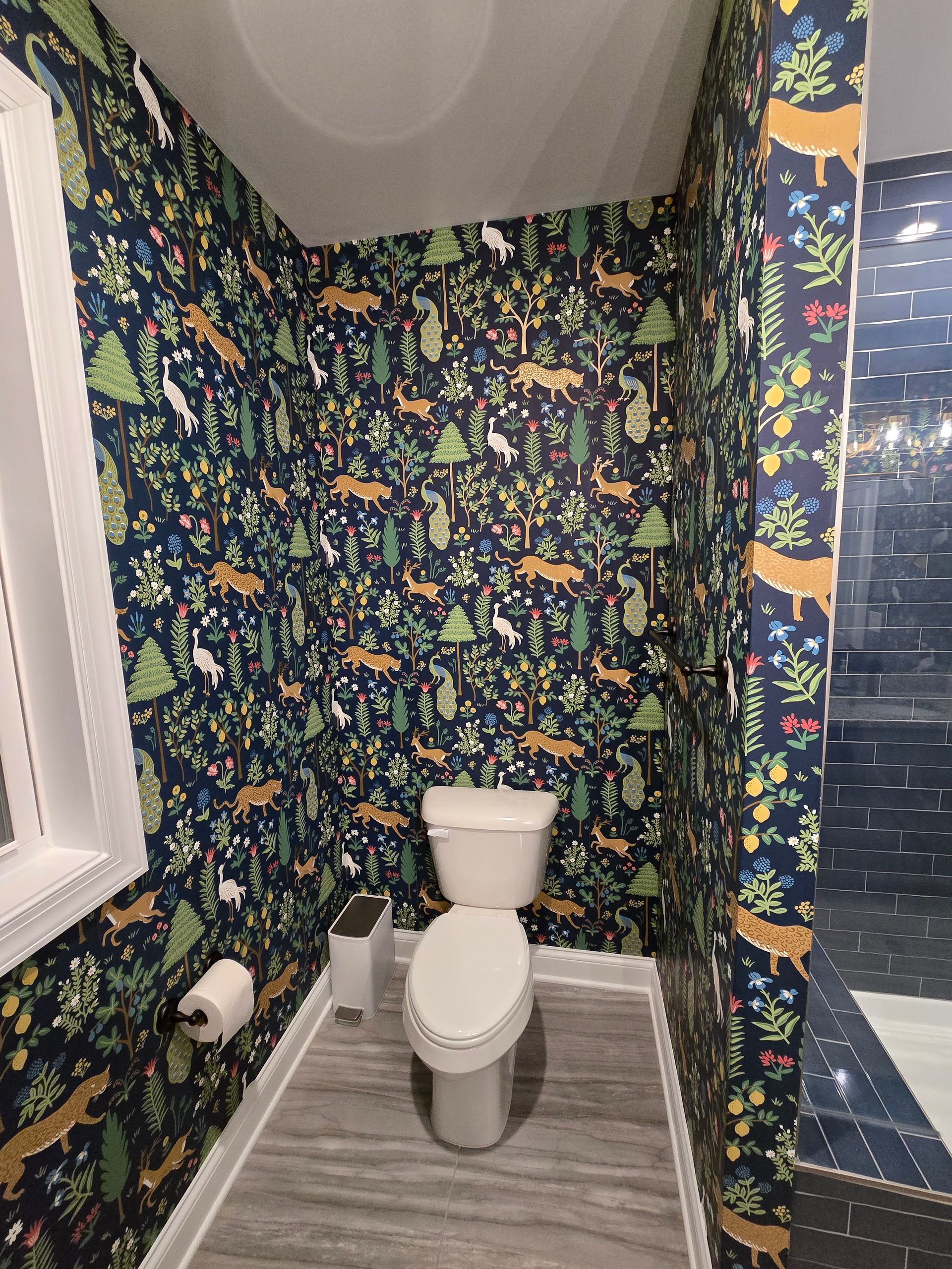 A small bathroom with forest-themed wallpaper, white toilet, and window, gray floor tiles.