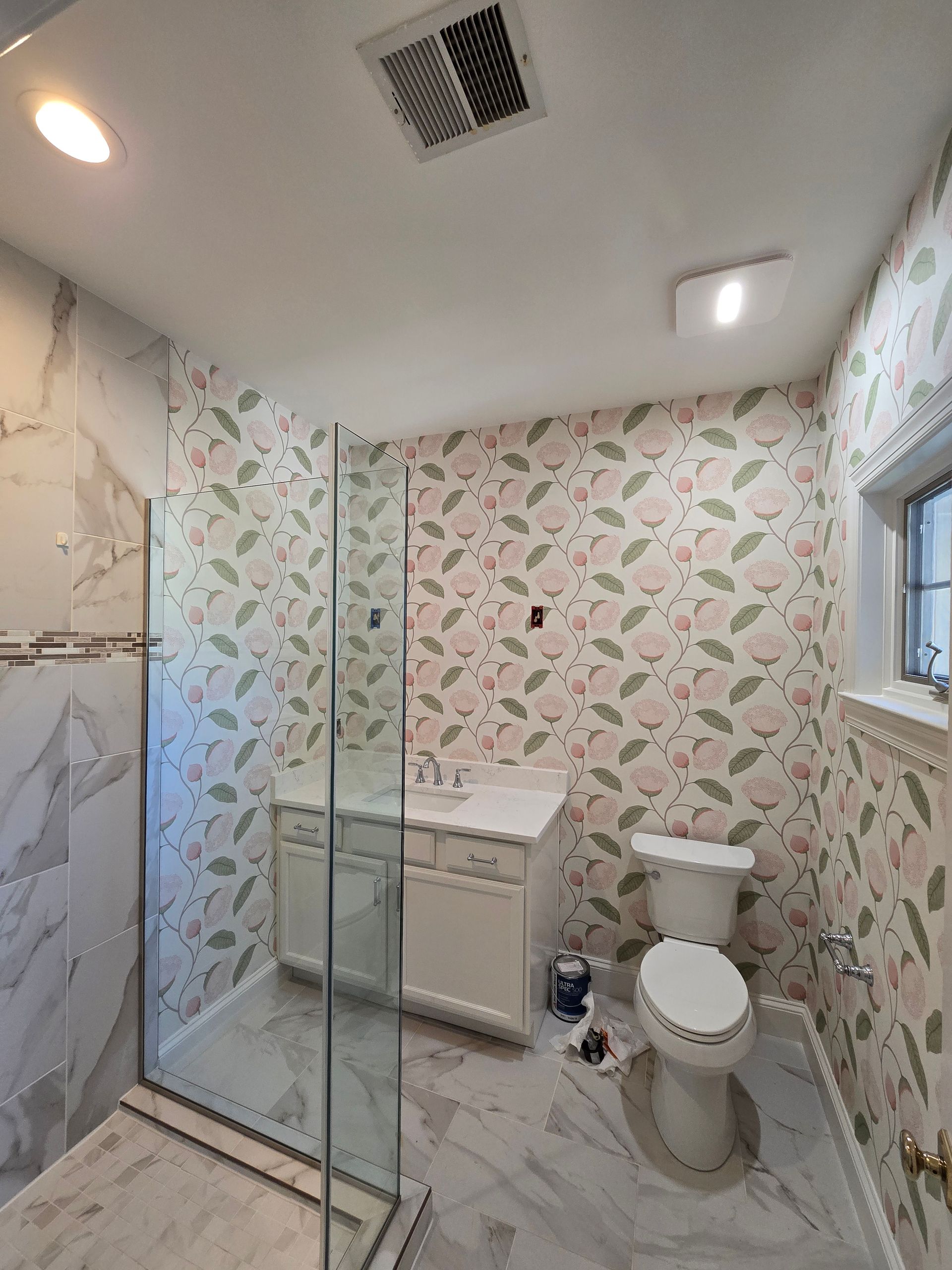 A bathroom with patterned floral wallpaper, a glass-enclosed shower, a white vanity, and a toilet on marble-look flooring.