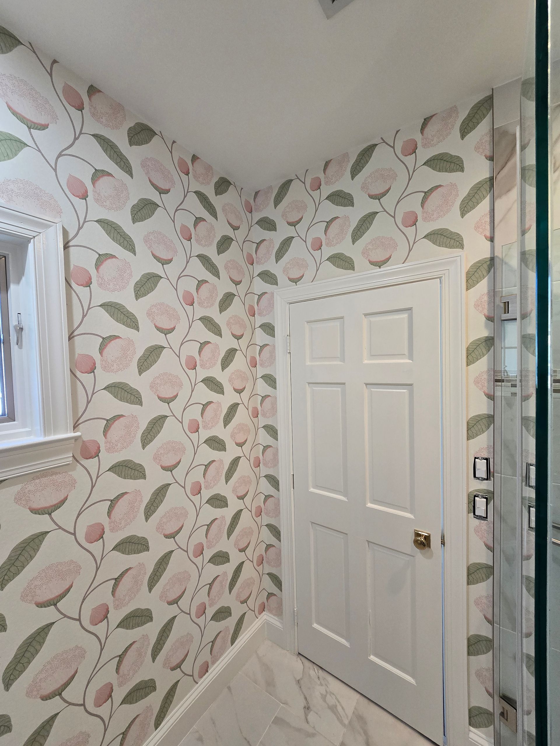 A corner in a room featuring white walls with pink floral wallpaper, a white door with decorative trim, and marble flooring.
