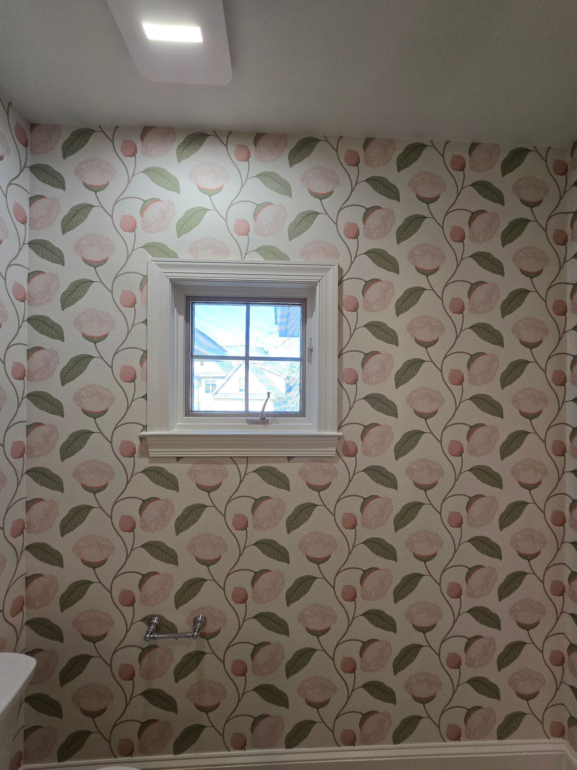 A bathroom wall featuring floral wallpaper with pink roses and green leaves, centered with a white-framed square window.