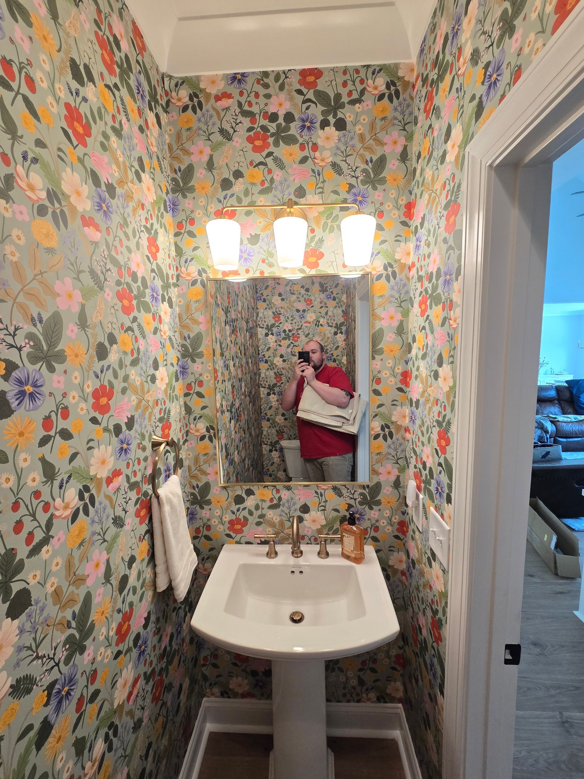 A person takes a selfie in a bathroom with vibrant floral wallpaper, a white pedestal sink, and a three-light fixture.
