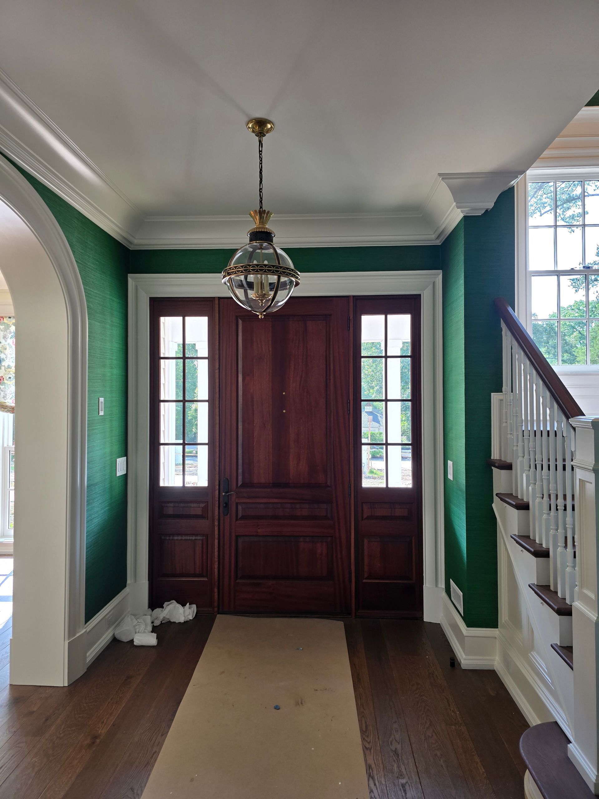 A wooden front door with side windows in an entryway with dark green walls, white trim, hardwood floors, and a pendant light.