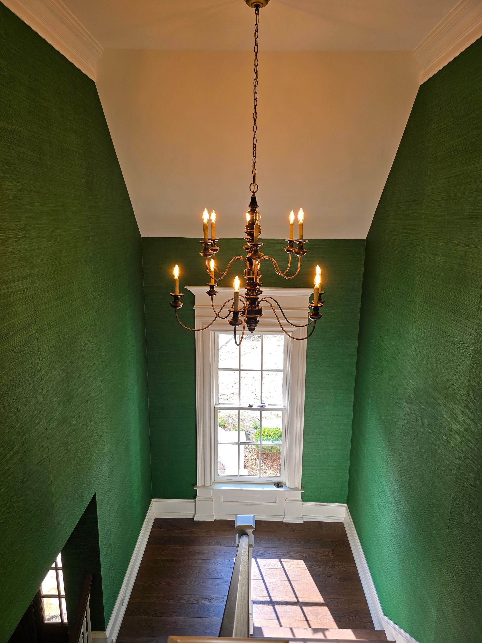 An elegant brass chandelier hangs in a tall stairwell with vibrant green textured walls and a bright window.