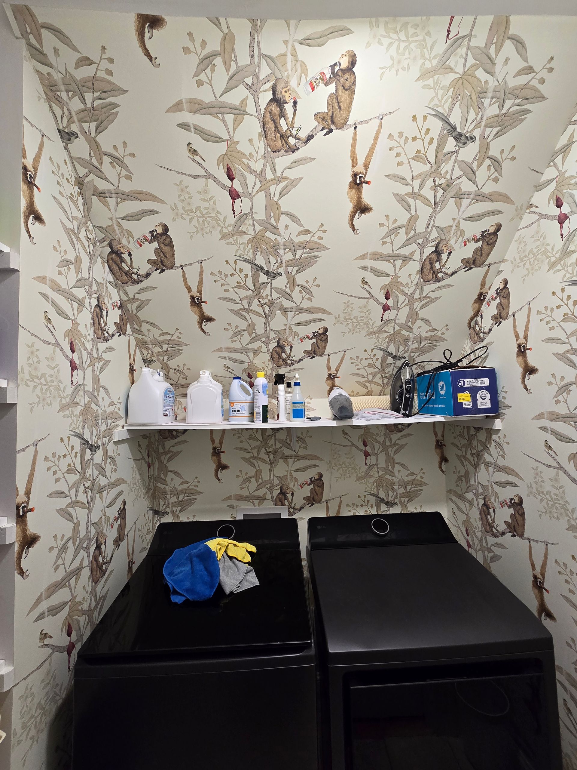 A laundry room with two black machines under a shelf, all set against wallpaper featuring a detailed jungle monkey pattern.