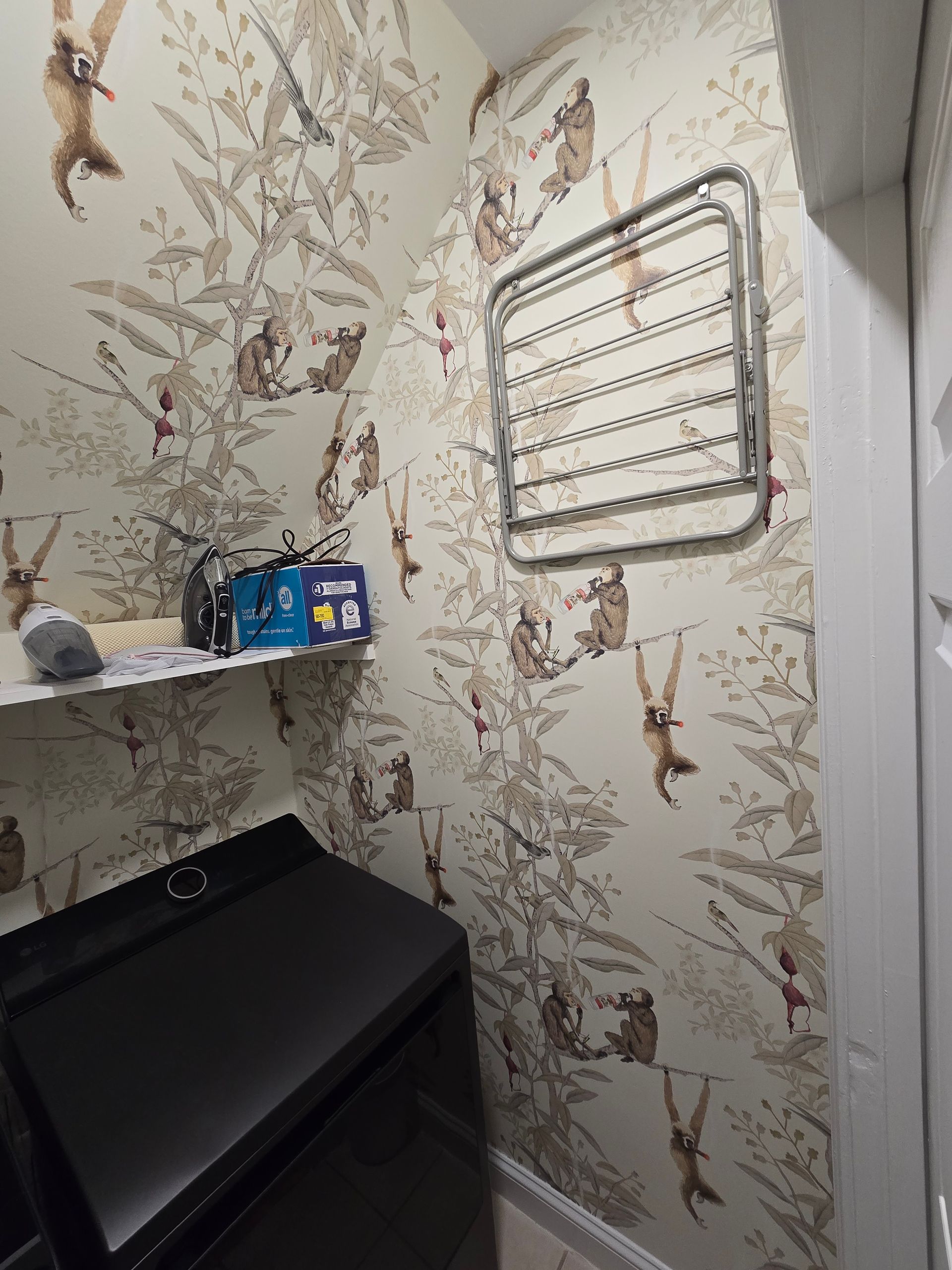 Laundry room featuring cream wallpaper with a whimsical monkey pattern, a black washing machine, and a wall drying rack.