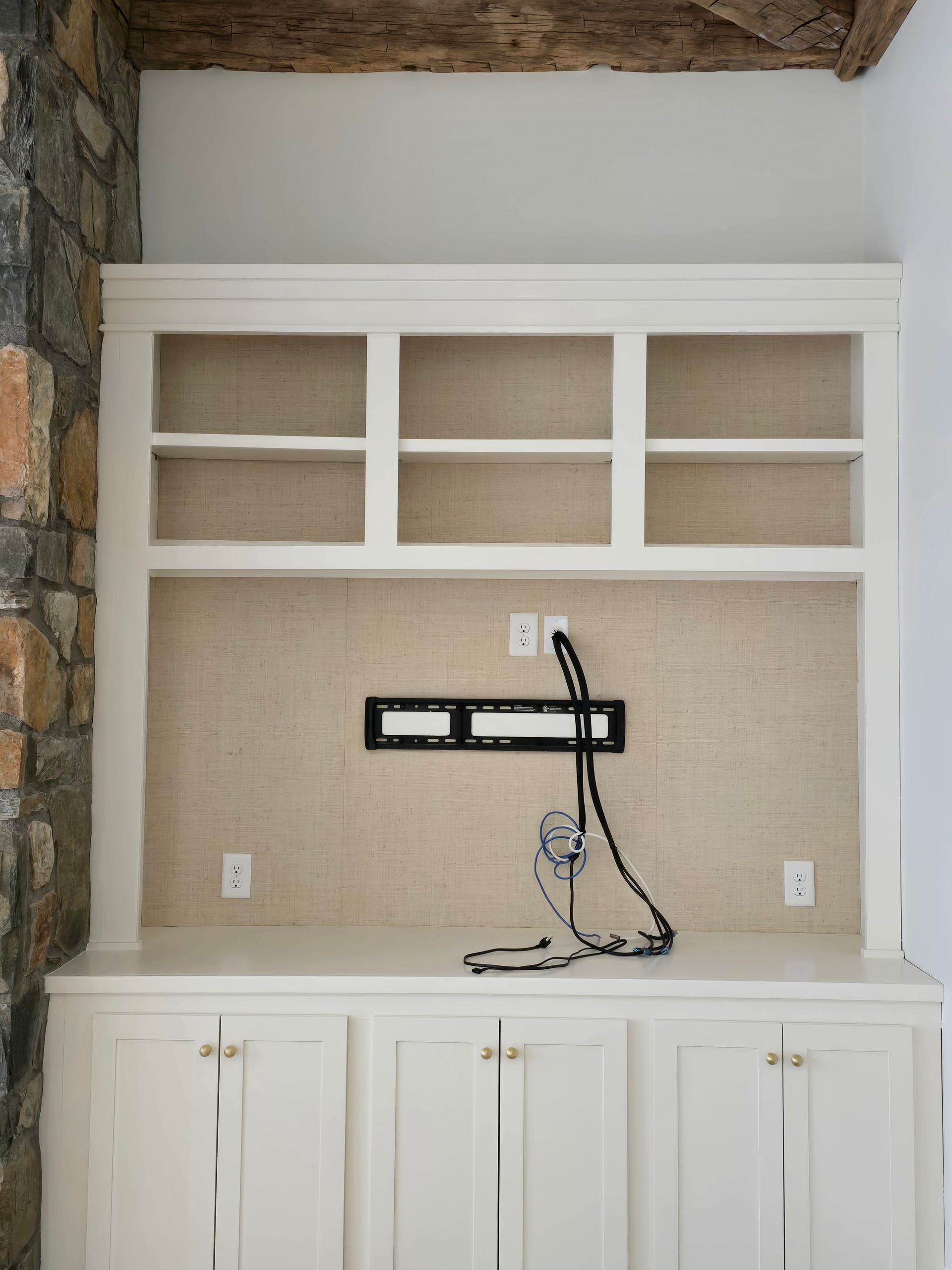A white custom entertainment center with upper shelving, a central television mount, and base cabinets against a stone wall.