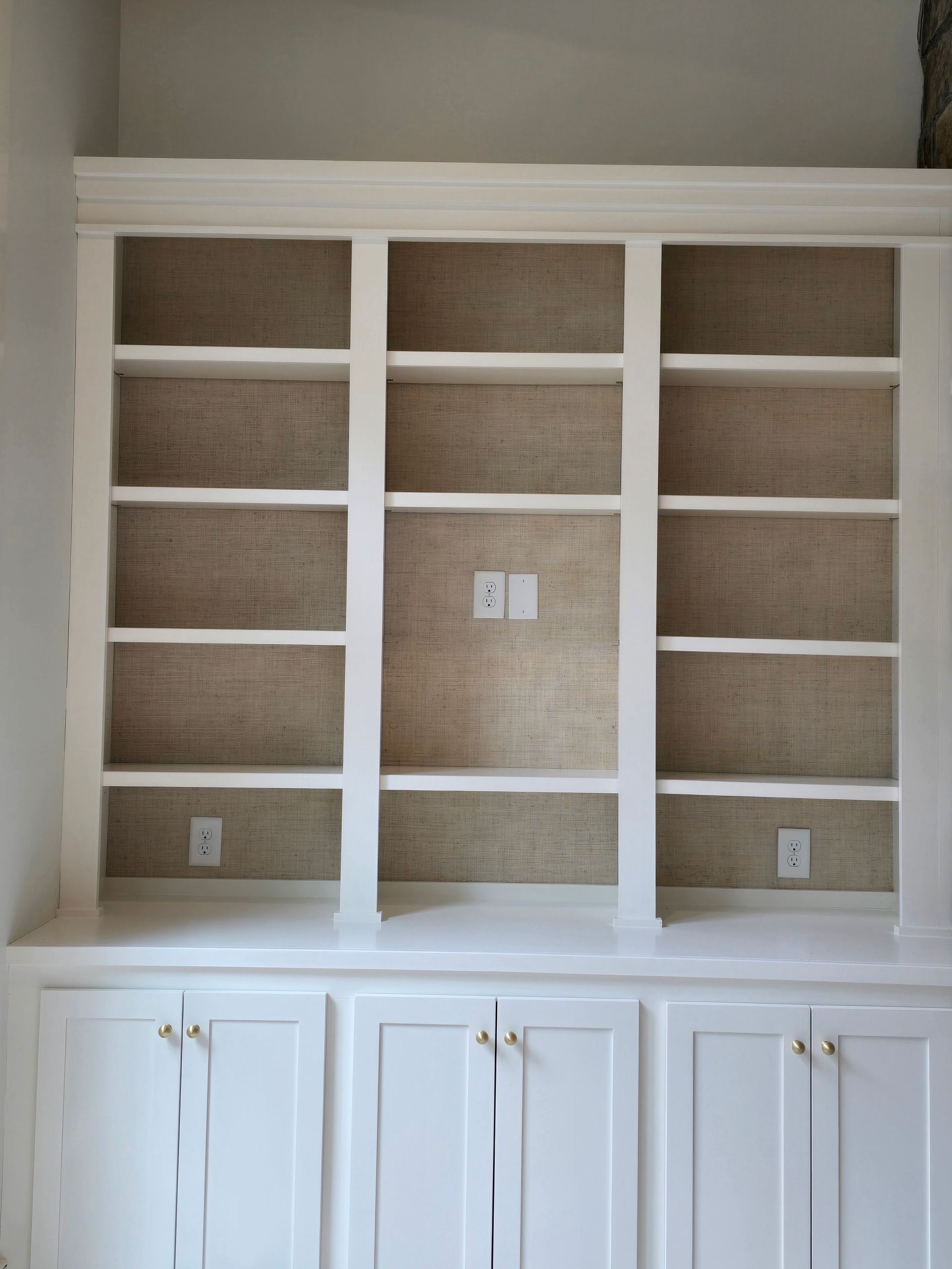 A built-in white bookshelf featuring three sections with textured, tan-colored back panels and integrated cabinetry below.
