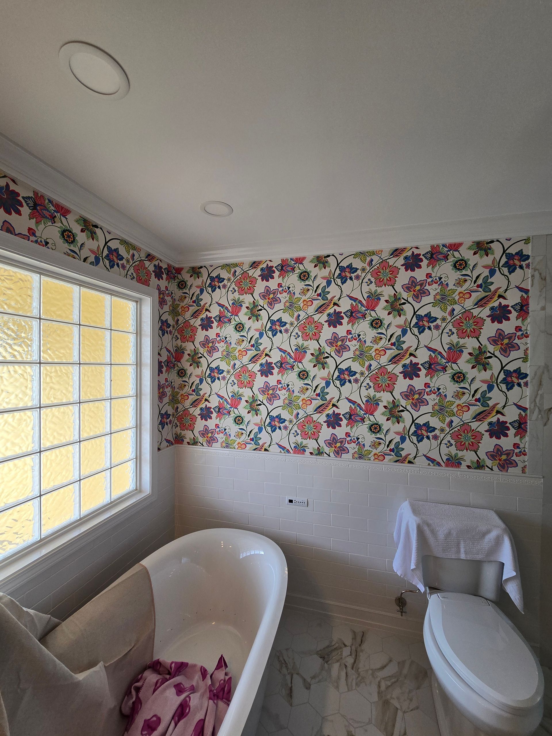 A bathroom with floral wallpaper above a white bathtub and toilet, next to a large window with grid-pane glass.