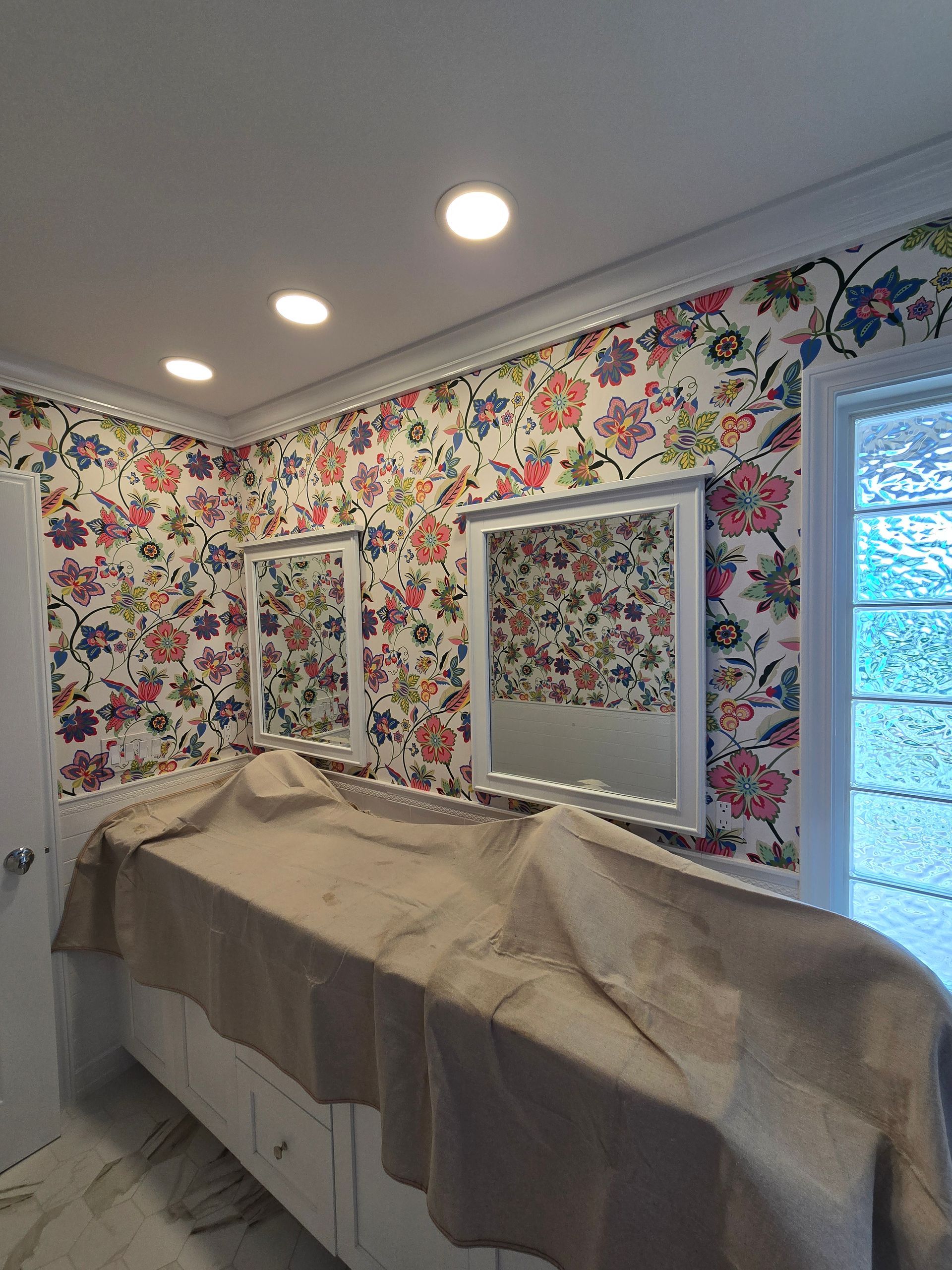 A bathroom vanity covered with a drop cloth, featuring mirrors against a wall with colorful floral wallpaper.