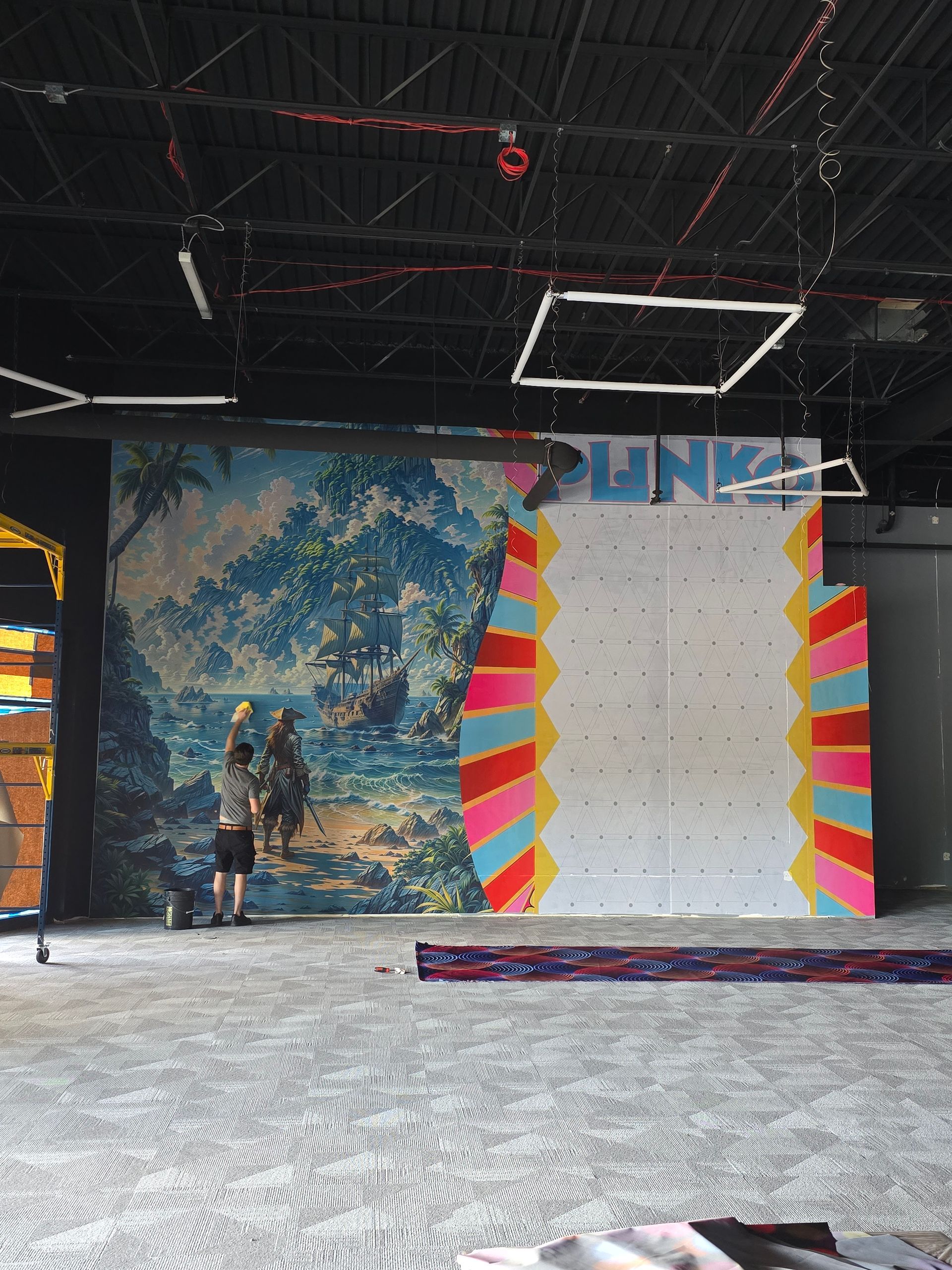 A person paints a tropical mural next to a climbing wall with colorful, striped borders in a large, industrial-style space.