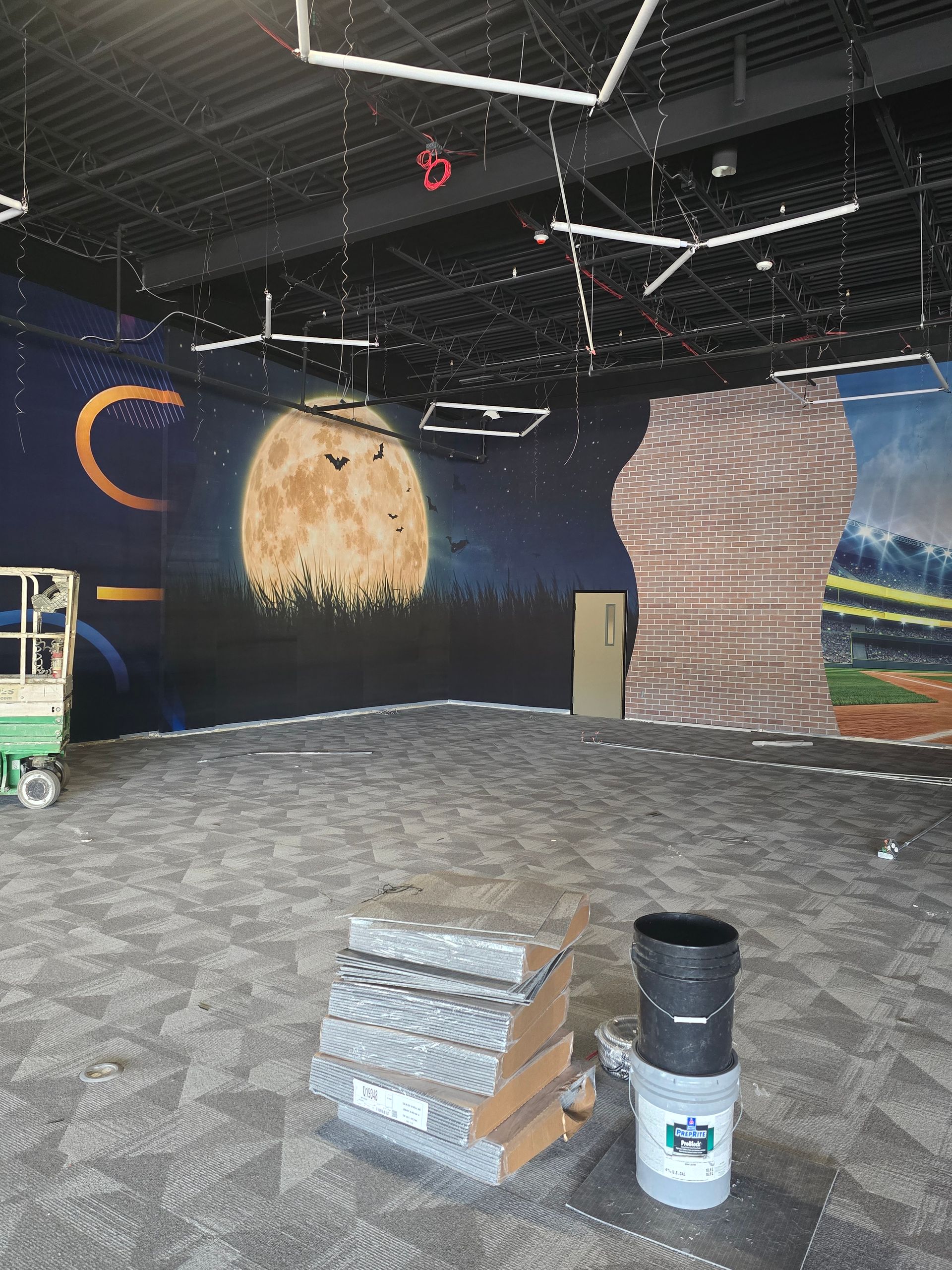 A room under construction with mural walls featuring a full moon, paint supplies on the floor, and a scissor lift.