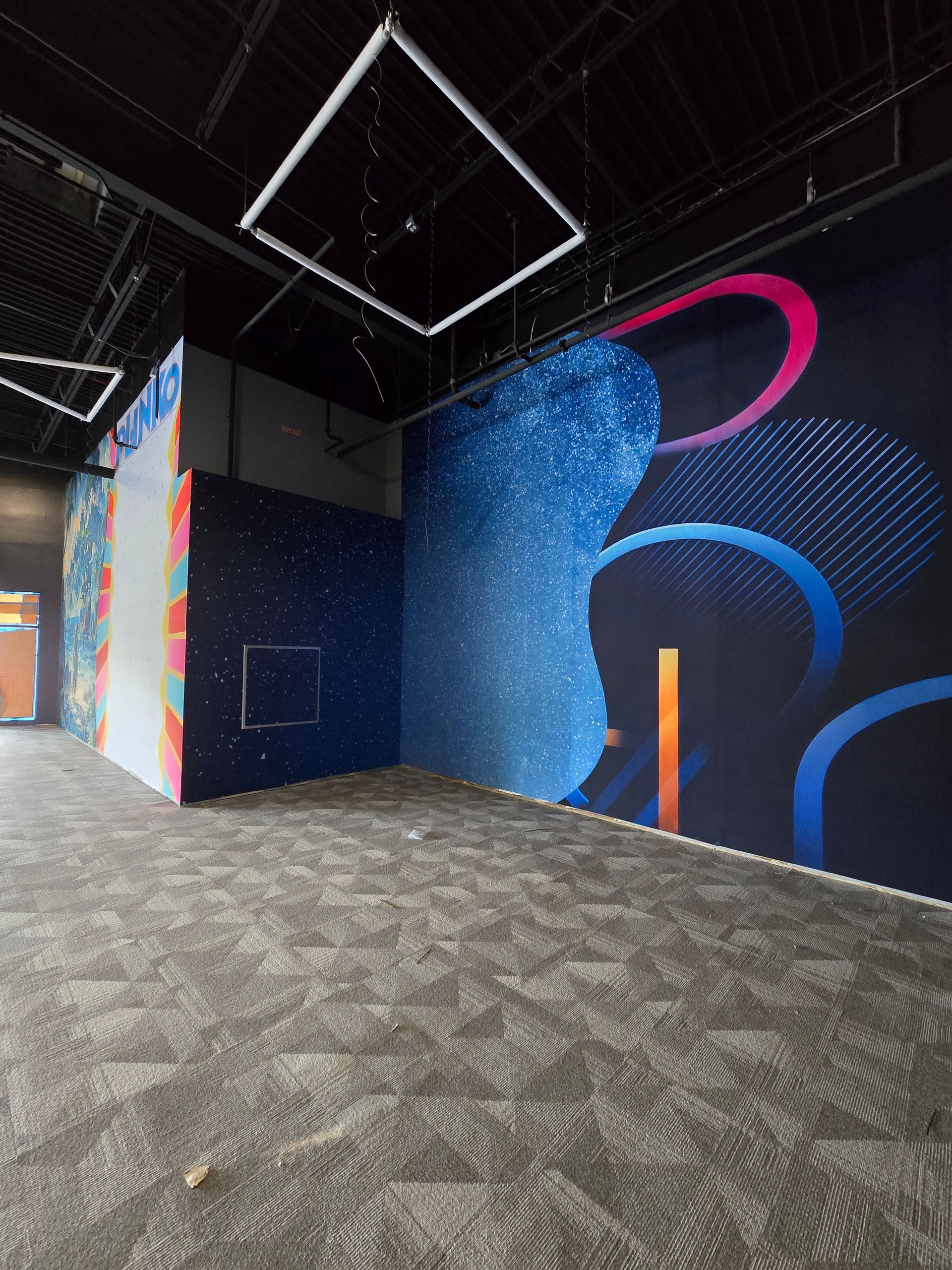 An indoor room with carpeted floor, featuring a large, colorful abstract mural with cosmic blue and pink patterns.