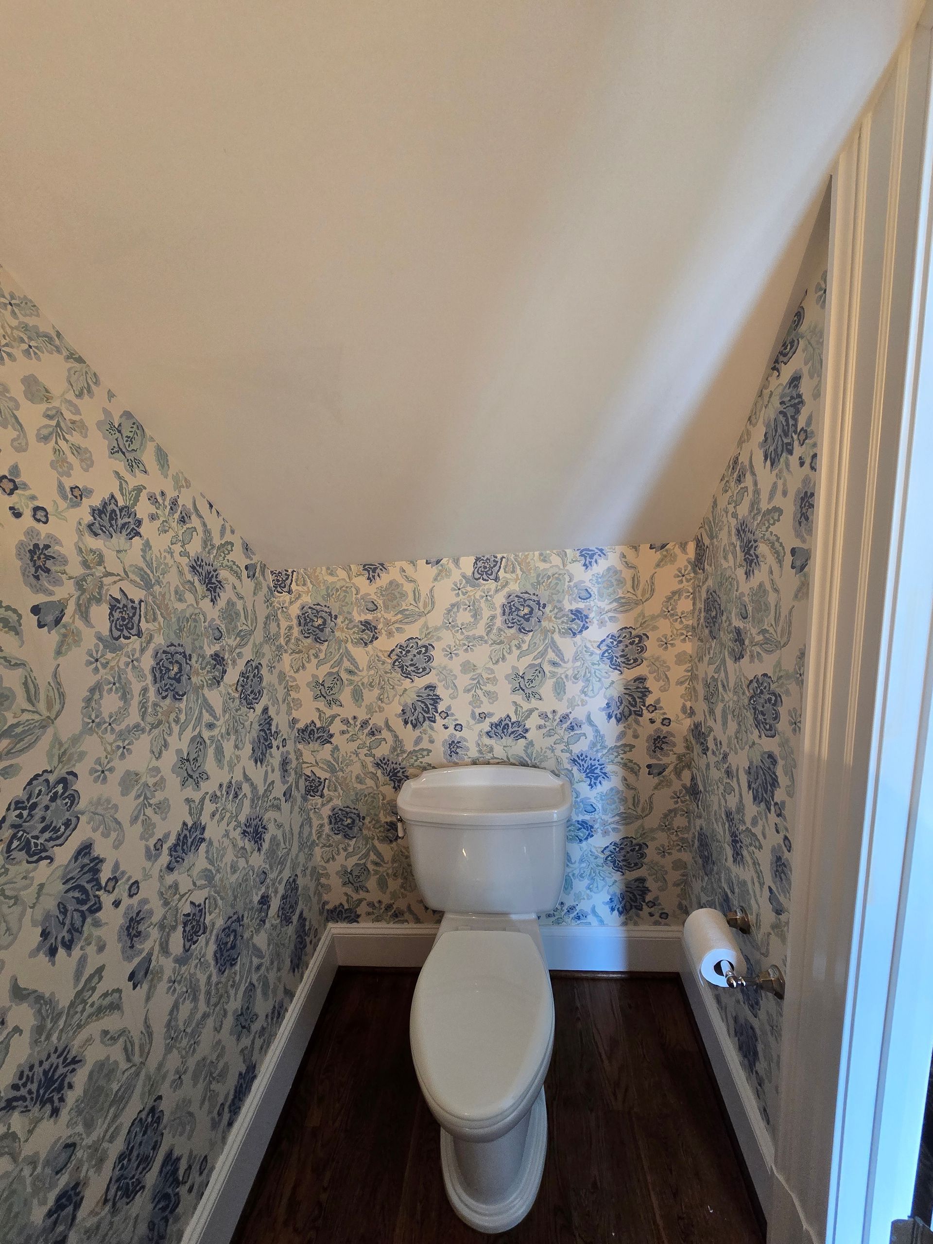 A white toilet in a small nook with slanted walls covered in blue and white floral wallpaper and dark wood flooring.