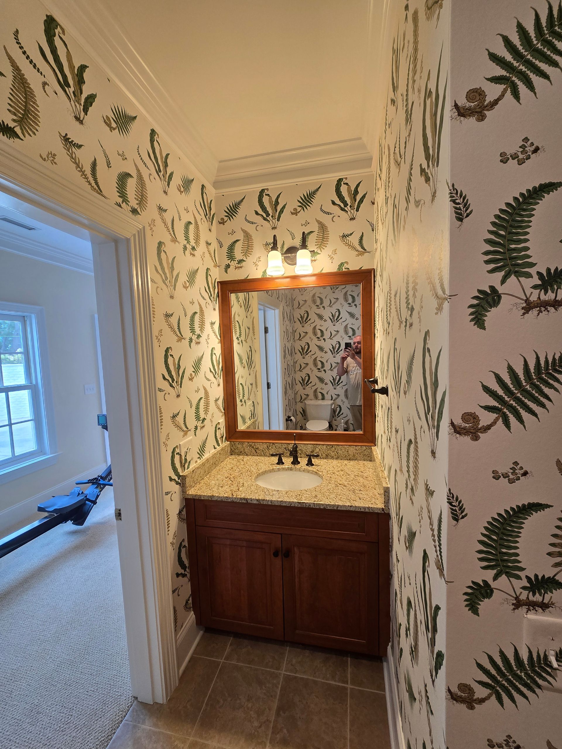 A small bathroom with white walls covered in a green fern wallpaper pattern, a wood vanity, and a framed mirror.