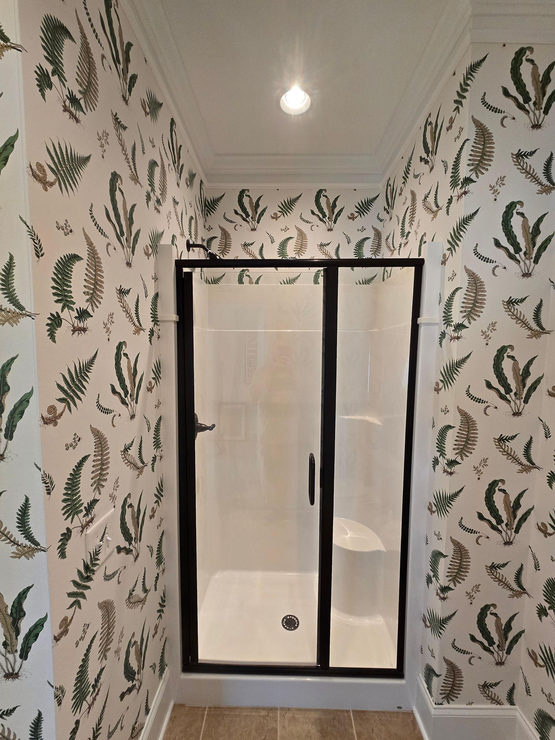A shower with a black frame surrounded by white walls featuring a repeating dark green fern leaf wallpaper pattern.