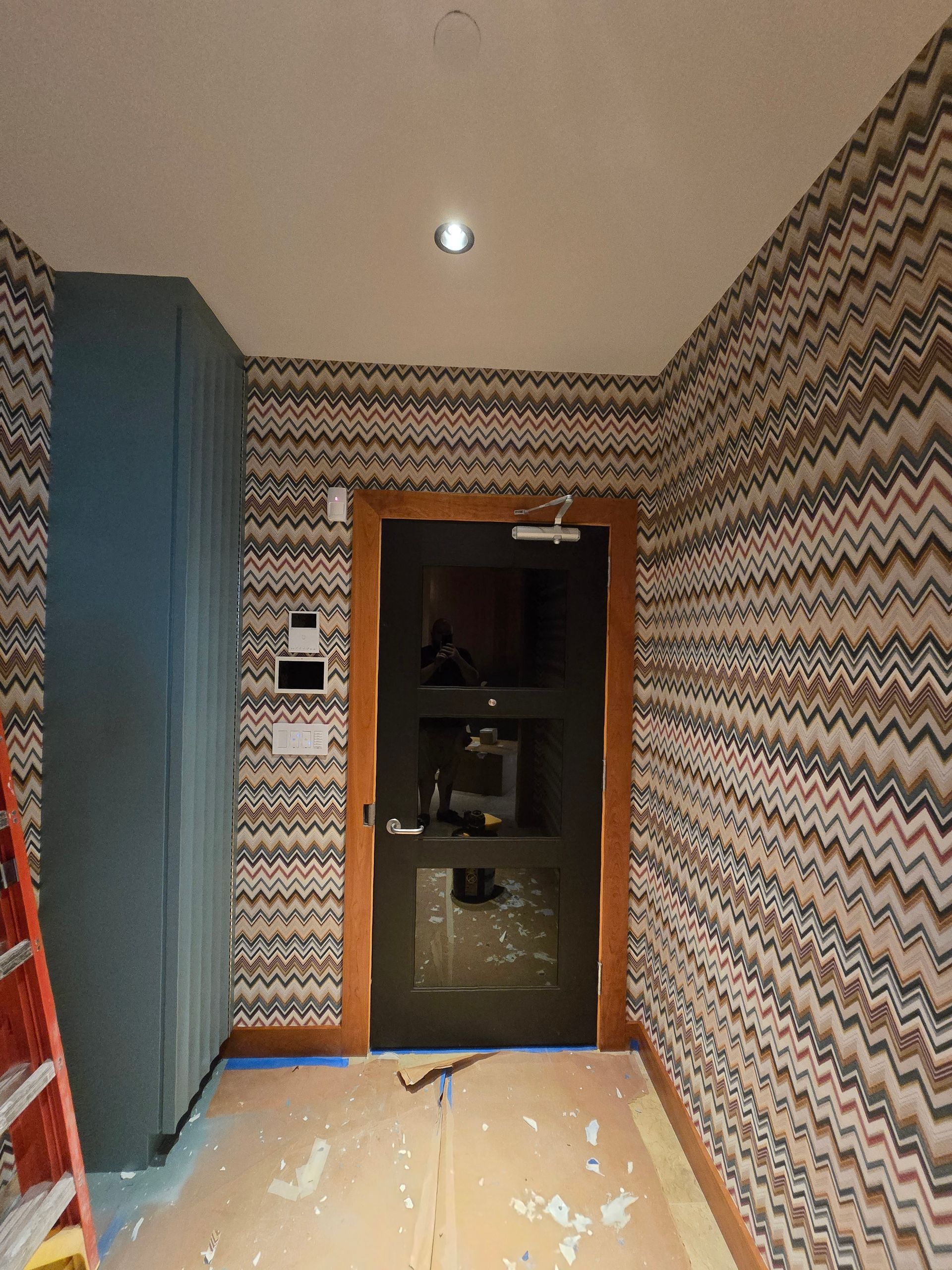 An interior view of a hallway featuring patterned zigzag wallpaper, a dark door, and protective paper on the floor.