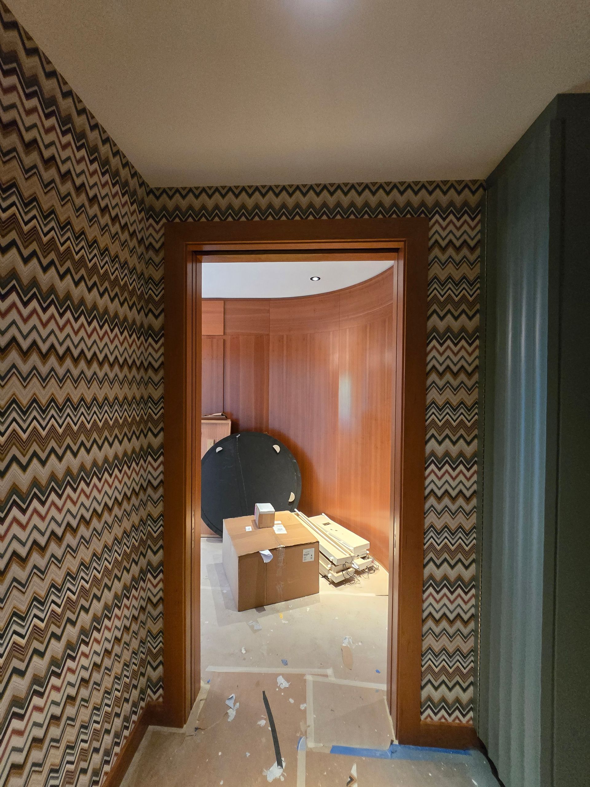A hallway with patterned chevron wallpaper leads into a room featuring wood-paneled walls, under construction.