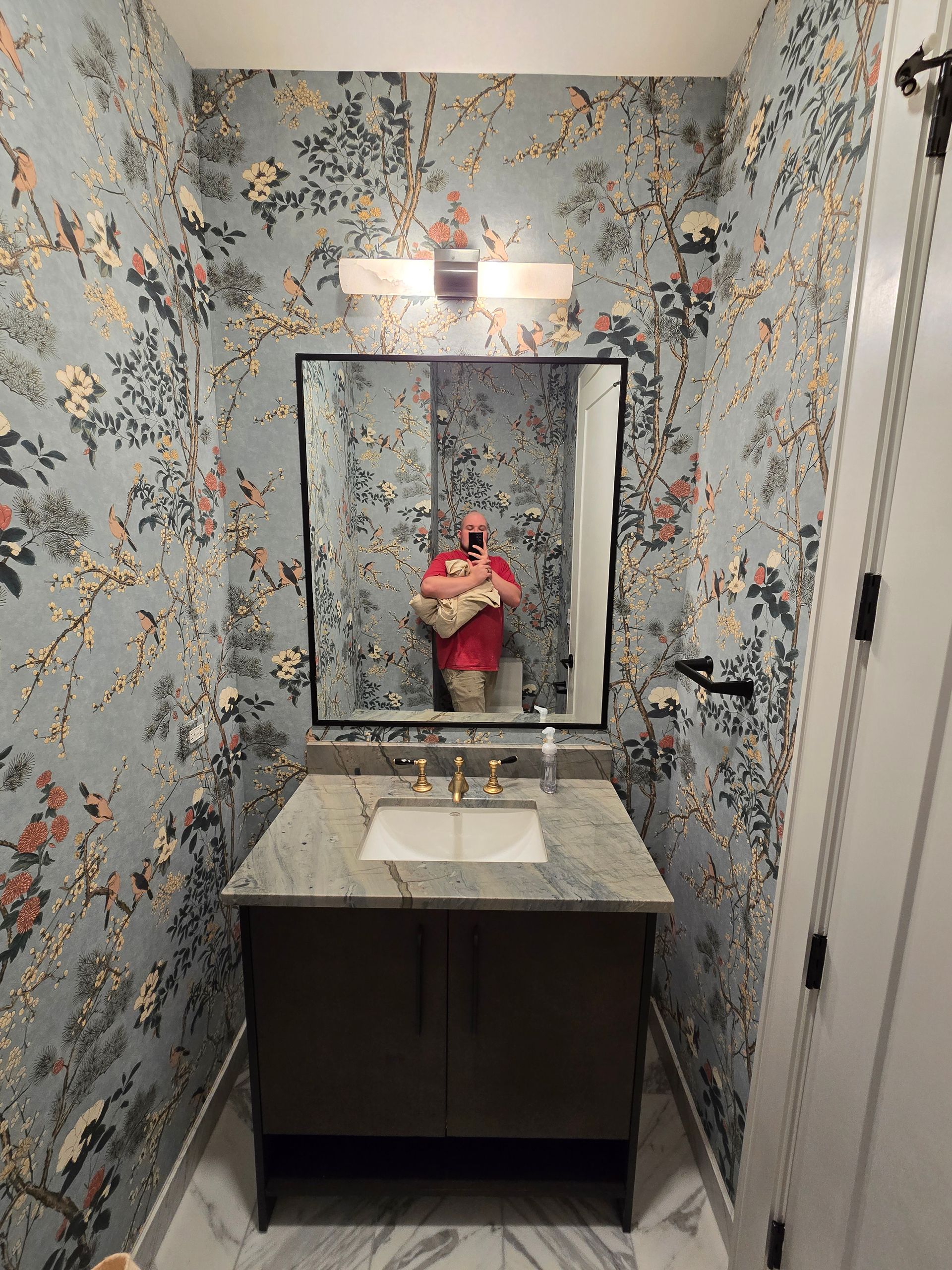 A powder room with blue floral wallpaper, a marble vanity, a rectangular mirror, and a person taking a photo in the mirror.