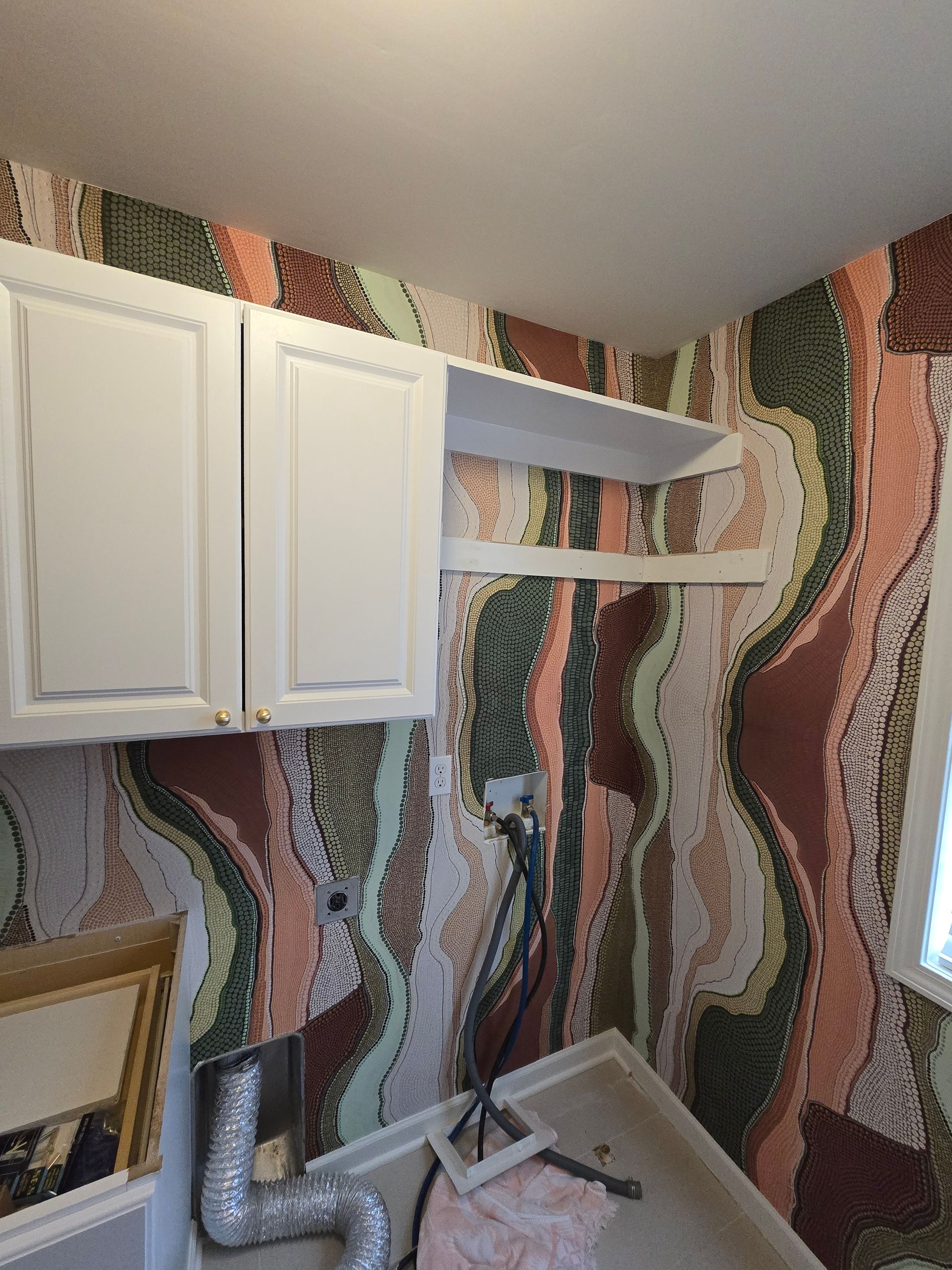 Laundry room walls covered in wavy, multicolored wallpaper patterns with white wall cabinets and exposed utility hoses.