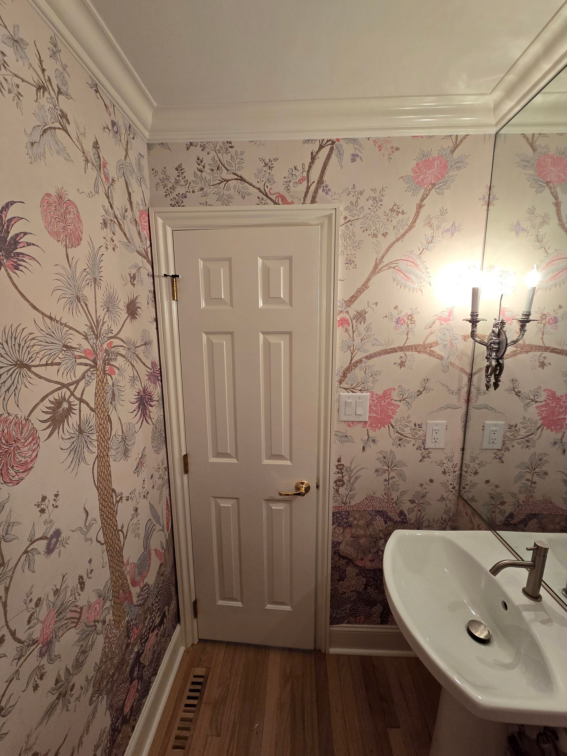 A bathroom with floral-patterned wallpaper, a white door, a wall-mounted light fixture, and a white pedestal sink.