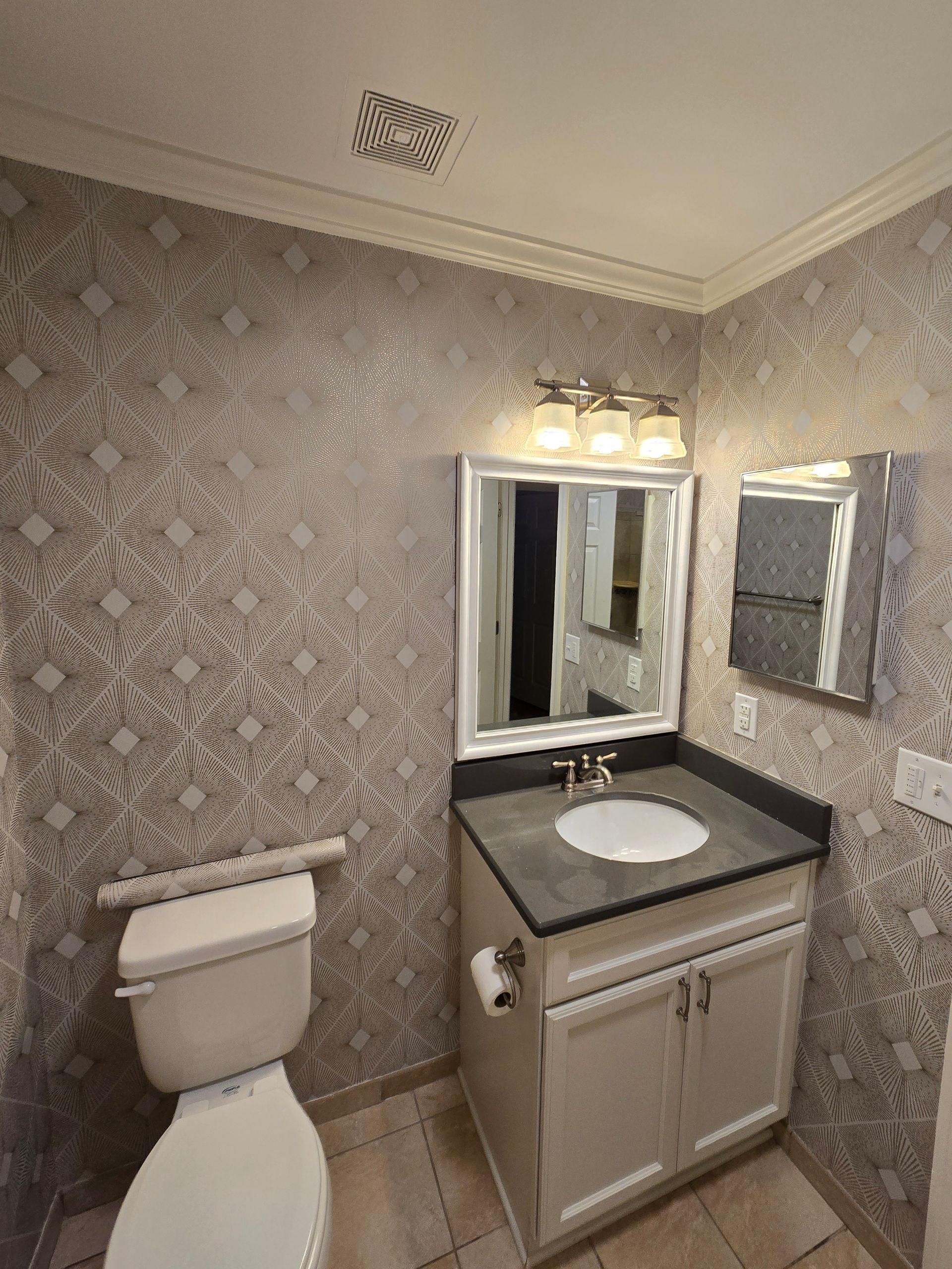 A powder room with patterned wallpaper, a light gray vanity with a dark countertop, a white toilet, and a vanity mirror.