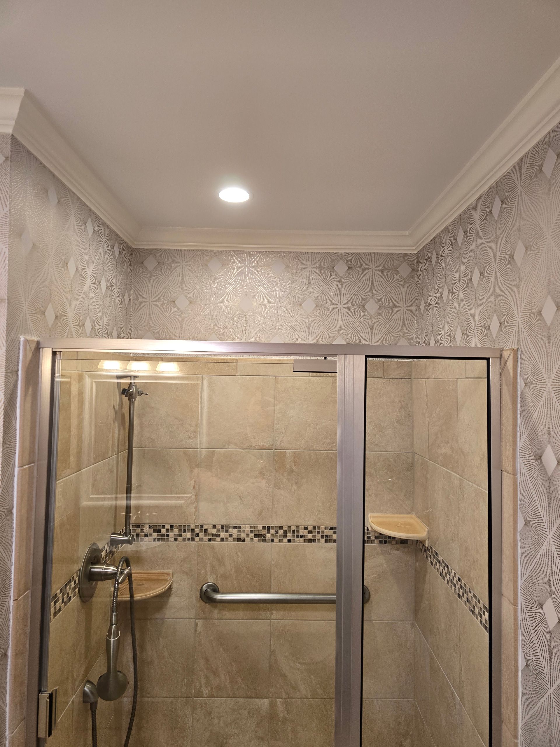 A glass-enclosed shower with beige tile walls, a grab bar, a handheld sprayer, and patterned gray wallpaper above.